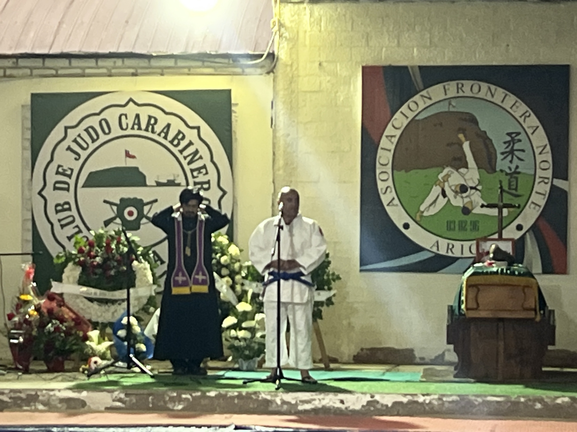 Two individuals are standing on a green carpeted stage in front of two large circular banners. The banner on the left is for "Club De Judo Carabineros" and depicts a green hill with a flag, a boat, and a circular emblem with a red dot. The banner on the right is for "Asociacion Frontera Norte" and shows a stylized depiction of two figures in judo uniforms grappling in front of a brown hill, with Japanese characters and the text "ARICA".
The person on the left is wearing a dark robe with purple and gold trim, resembling clerical attire. They have their hands raised to their head. The person on the right is wearing a white judogi with a blue belt and is standing with their hands clasped in front of them. Both individuals are positioned in front of floral arrangements.
To the right of the stage, there is a wooden structure topped with a dark cloth, resembling a podium or altar with a wooden box on top. The scene appears to be outdoors, with a brick wall in the background and a light fixture visible at the top. The location is identified as Arica, Chile.