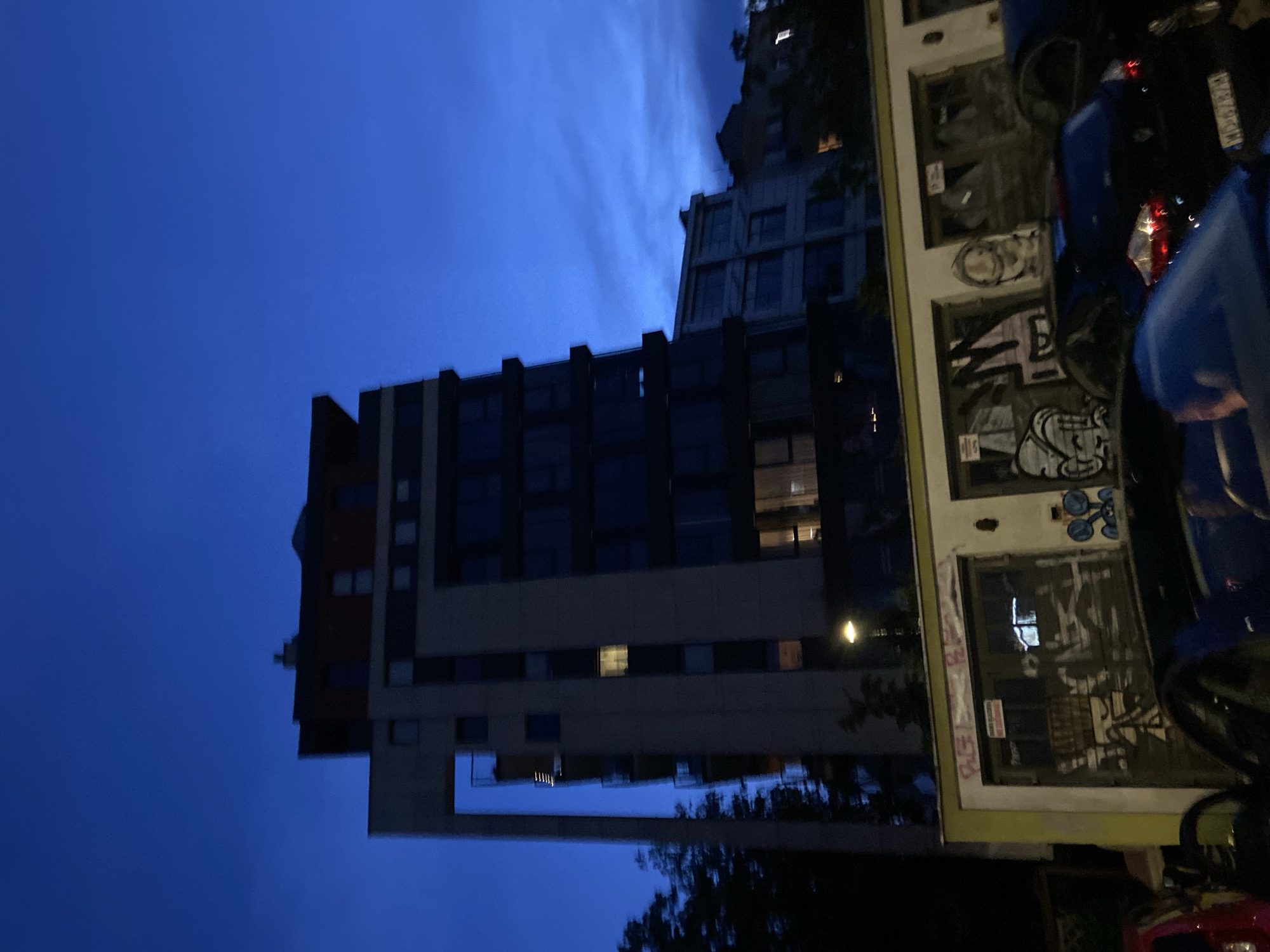 The image depicts a street scene in Warsaw, Poland, at dusk or night. A modern, multi-story building with balconies and darkened windows dominates the background, its upper floors featuring a darker, reddish-brown section. In the foreground, a row of garage doors is visible, heavily adorned with graffiti. Several cars are parked in front of the garages, including a blue hatchback with its license plate partially obscured. The sky is a deep blue, with some wispy clouds. A streetlamp illuminates the scene from the right side of the image. The overall atmosphere is urban and nocturnal.