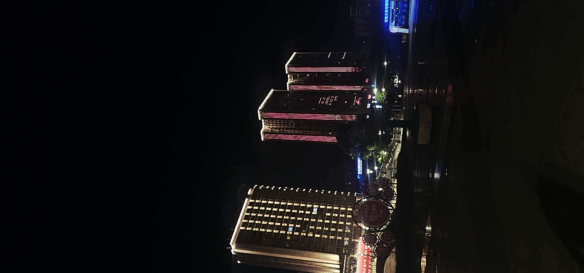 This is a nighttime, low-angle photograph taken in Beibei, China. The image captures two illuminated high-rise buildings in the background. The building on the left is lit with yellow lights, showcasing a grid pattern of illuminated windows. The building on the right is illuminated with vertical red lights. In the foreground, there are circular decorative structures with Chinese characters on them, some of which are illuminated. The ground appears wet, reflecting the lights from the buildings and decorative elements, suggesting it may have recently rained. The overall scene is dark with artificial lighting from the buildings and decorative elements providing the primary illumination.