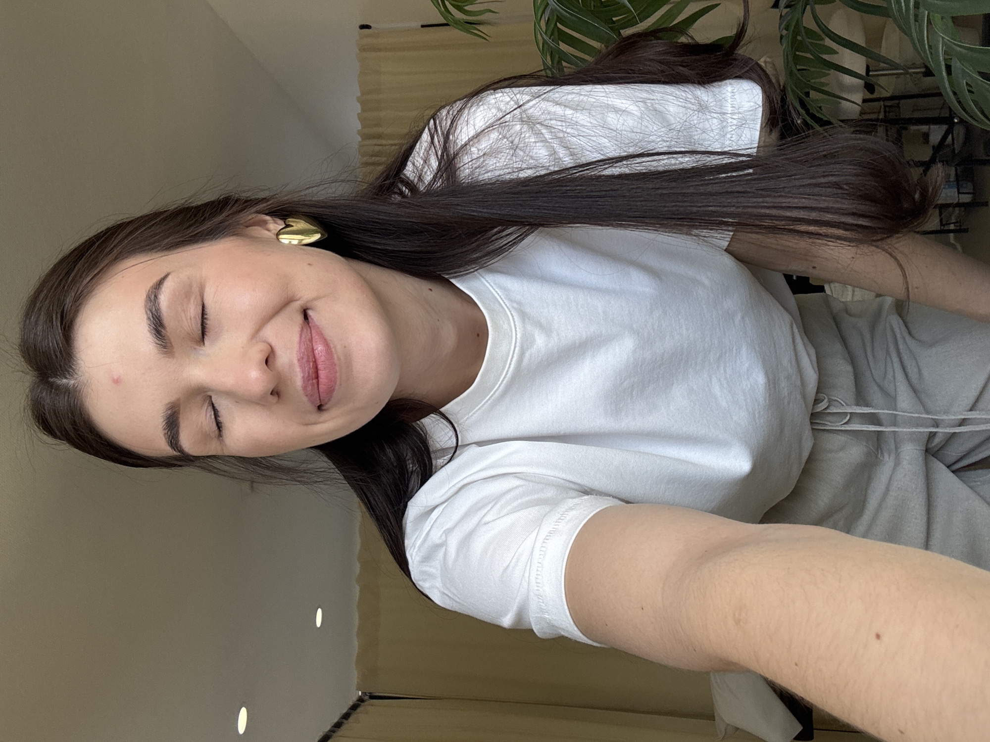 An indoor, eye-level shot features a female subject in a casual pose. She has long, dark brown hair, fair skin, and her eyes are closed with a slight smile on her lips. A small reddish blemish is visible on her forehead. She wears a plain white short-sleeved t-shirt and light gray drawstring sweatpants. Gold-colored heart-shaped stud earrings are visible on her ears. Her left arm extends into the lower left foreground, suggesting she is holding the camera. The background consists of light beige or tan curtains on the right and a darker vertical partition on the far left. A large green leafy plant is situated in the lower right background. The ceiling is light-colored with two visible recessed circular lights. Unidentifiable dark shapes are present in the lower center background.