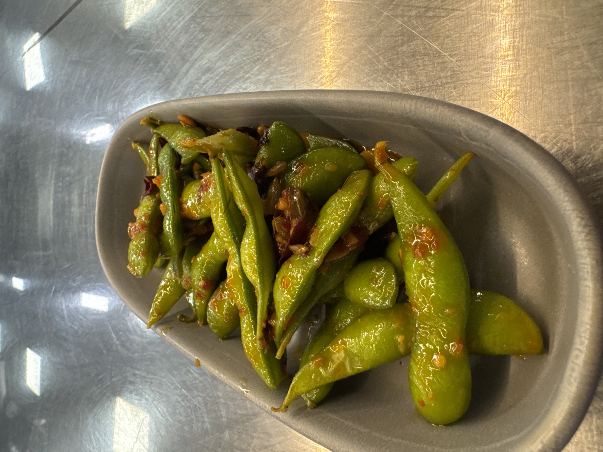 A serving of bright green edamame pods, likely seasoned, is presented in a modern, oval-shaped gray bowl. The edamame pods exhibit a glossy sheen, indicating they are coated in a sauce or oil, which also contains visible reddish-orange specks, possibly chili flakes or other spices. The pods are arranged loosely within the bowl, showcasing their vibrant color and texture.
The bowl itself is a matte, light gray or stone-colored ceramic, featuring a simple, slightly raised rim. It rests on a highly reflective, brushed metallic surface, most likely stainless steel. This surface is extensively covered with linear scratches and scuff marks, characteristic of a commercial kitchen counter or food preparation area.
The metallic surface prominently reflects multiple distinct, rectangular sources of overhead artificial light, creating bright highlights and emphasizing the industrial nature of the setting. The overall scene is brightly illuminated, suggesting an indoor environment. There are no people present, and no activity is occurring beyond the static presentation of the dish. No visible text appears in the image.