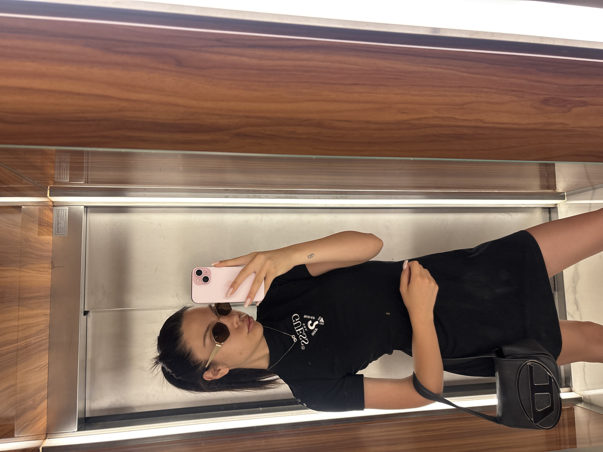 In Sofia, Bulgaria, a young woman with her hair in a ponytail stands in front of a mirror in what appears to be an elevator or a well-lit hallway with wood-paneled walls. She is taking a selfie with her pink smartphone, holding it in her right hand. She is wearing a black t-shirt with a "Guess Jeans" logo on it, a black skirt, sunglasses, and a silver necklace. A black handbag with a silver "Diesel" logo is slung over her left shoulder. Her left arm is resting on her skirt. The background features a reflective surface, likely a mirror, which shows a portion of the environment behind her. The overall impression is a stylish and modern urban setting.