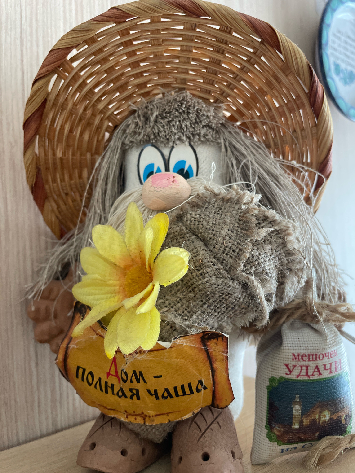 The image shows a close-up of a decorative doll wearing a straw hat and holding a yellow flower. The doll has large blue eyes and a pink nose. It is dressed in burlap clothing. A small sachet with Russian writing and an image of a lighthouse is attached to its side. There is also a sign with Russian writing that says "Дом - Полная Чаша" which translates to "Home - Full Cup". The doll is placed on a wooden surface. The background consists of a light-colored wall. The image does not depict the city of Pattaya City, Thailand.