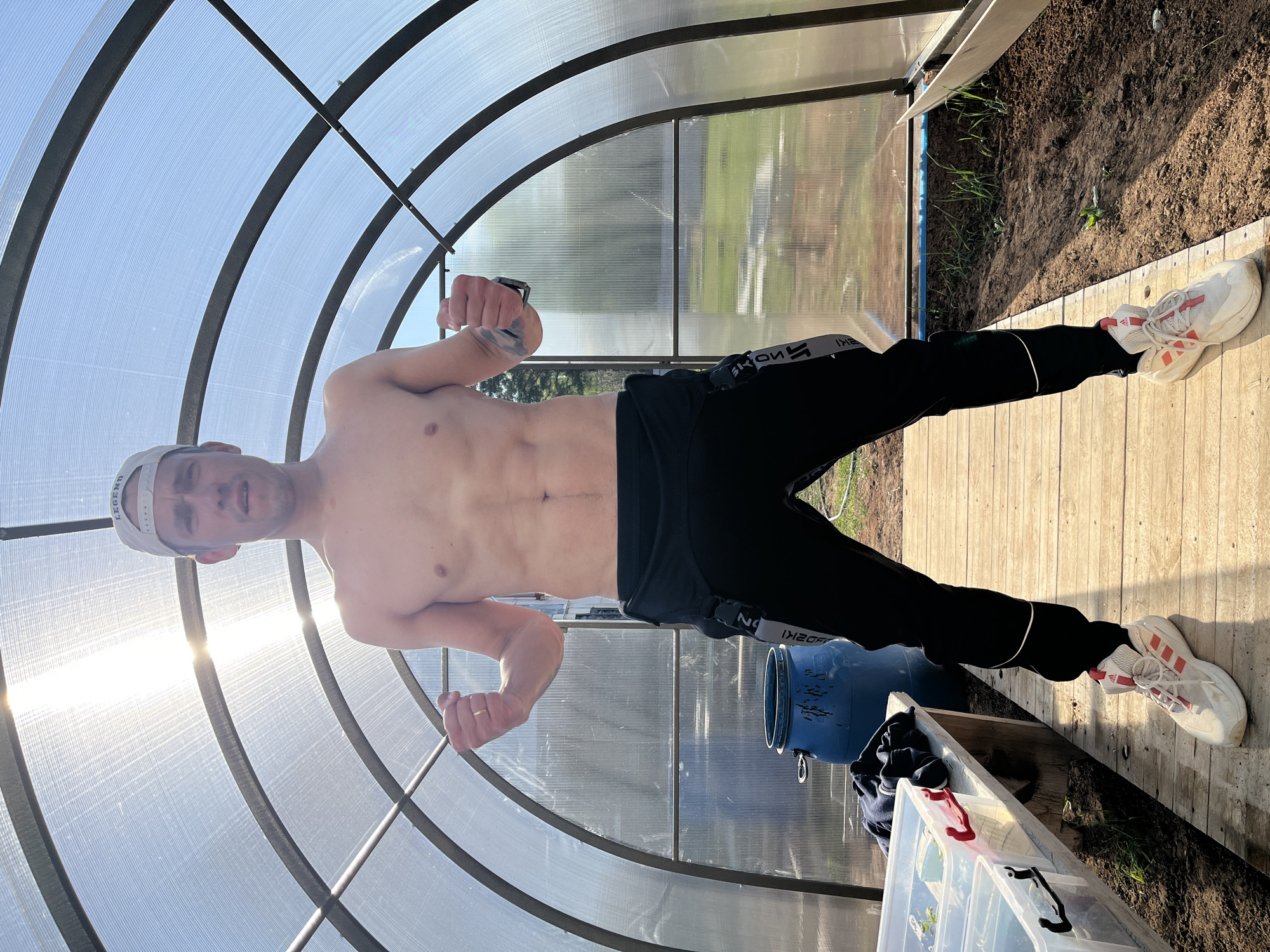 A shirtless man stands in a greenhouse. He is wearing black pants, a white cap, and white sneakers with red stripes. The man is flexing his arms. Behind him is a clear blue water barrel and some gardening supplies. The greenhouse has a plastic roof and a wooden floor, with dirt on either side. The location is consistent with a suburban area such as Chaikovsky, Russia.