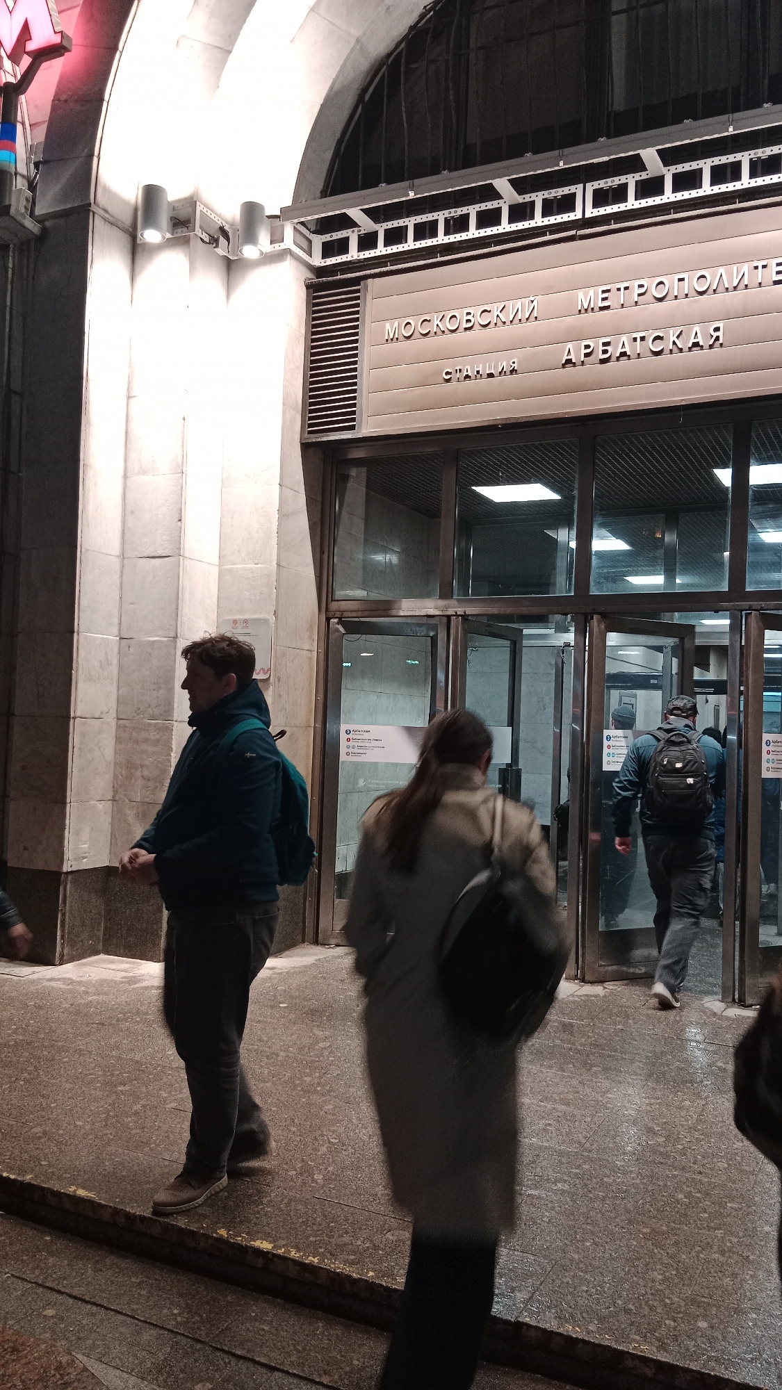 The image shows the entrance to the Arbatskaya station of the Moscow Metro in Moscow, Russia. The station entrance is marked by a large sign in Russian script that reads "Moskovskiy Metropoliten" and "Arbatskaya." Three people are visible. A man with a blue jacket and green backpack is standing on the left, while a woman with long hair wearing a grey coat and carrying a backpack is walking toward the doors. Another man wearing a backpack is entering the station. The entrance is made of glass doors and a stone archway with metal accents. The lighting is a mix of natural light and artificial light from fixtures above the entrance and within the station. The ground is a speckled stone.