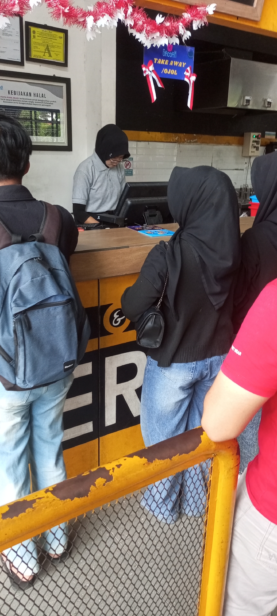 The image shows a counter area with at least three people. One person, wearing a gray polo shirt and a black hijab, stands behind the counter and appears to be operating a computer terminal. Another person, also wearing a black hijab, stands in front of the counter, facing away from the camera, and is wearing a black jacket and blue jeans. A third person, partially visible on the left side of the image, wears a black jacket and blue jeans and has a blue backpack. Behind the counter, a sign reads "GACOAN TAKE AWAY / OJOL". The counter itself is yellow and black, with a large white "ER" logo visible on the lower section. A yellow metal railing with a chain-link fence is in the foreground. The overall environment suggests a food service establishment, possibly a restaurant or cafe. The context of "Dapur, Indonesia" indicates the geographical location.