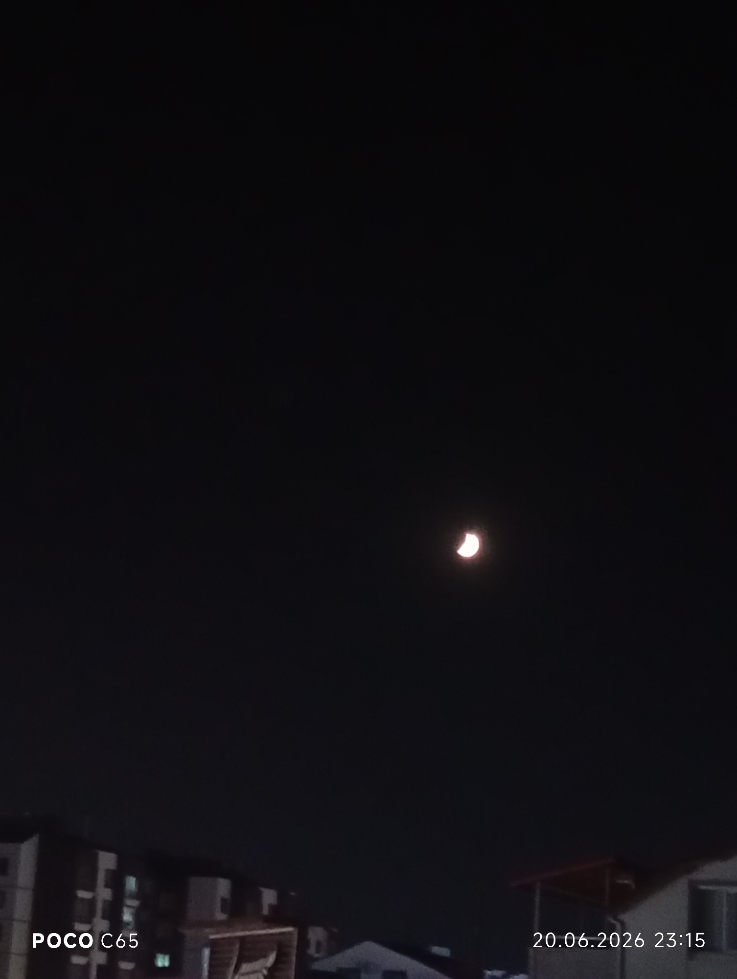 The image shows a nighttime view of a crescent moon in a dark sky over the city of Körfez, Turkey. In the lower portion of the image, the outlines of buildings and rooftops are visible. The text "POCO C65" is overlaid on the bottom left, and "20.06.2026 23:15" is overlaid on the bottom right, indicating the device used and the date and time the photo was taken. There are no people visible in the image. The overall scene is dark and illuminated only by the faint light of the moon and some artificial lights from the buildings.