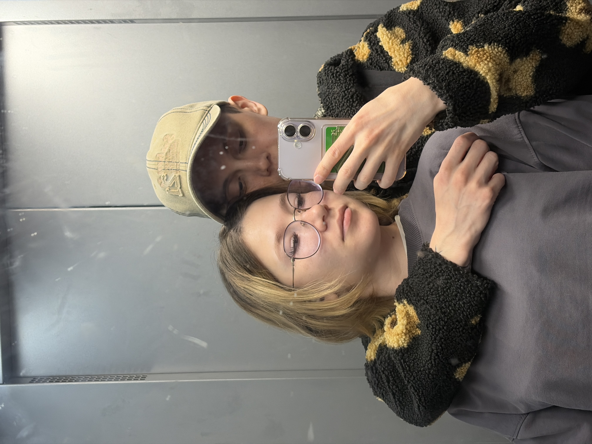 This is a reflection selfie taken by two people in an elevator. The person in the foreground is wearing glasses and has blonde hair. The person in the background is wearing a baseball cap and a fuzzy jacket with a leopard print pattern. They are both looking at the camera, and the person in the background is holding a smartphone to take the picture. The elevator appears to be modern, with gray metallic walls and a grated ventilation system. There are some smudges on the mirror, which are visible in the reflection. The lighting is bright, suggesting it is daytime. The overall mood of the image is casual and intimate, with the two people appearing close to each other. No visible text is present in the image.