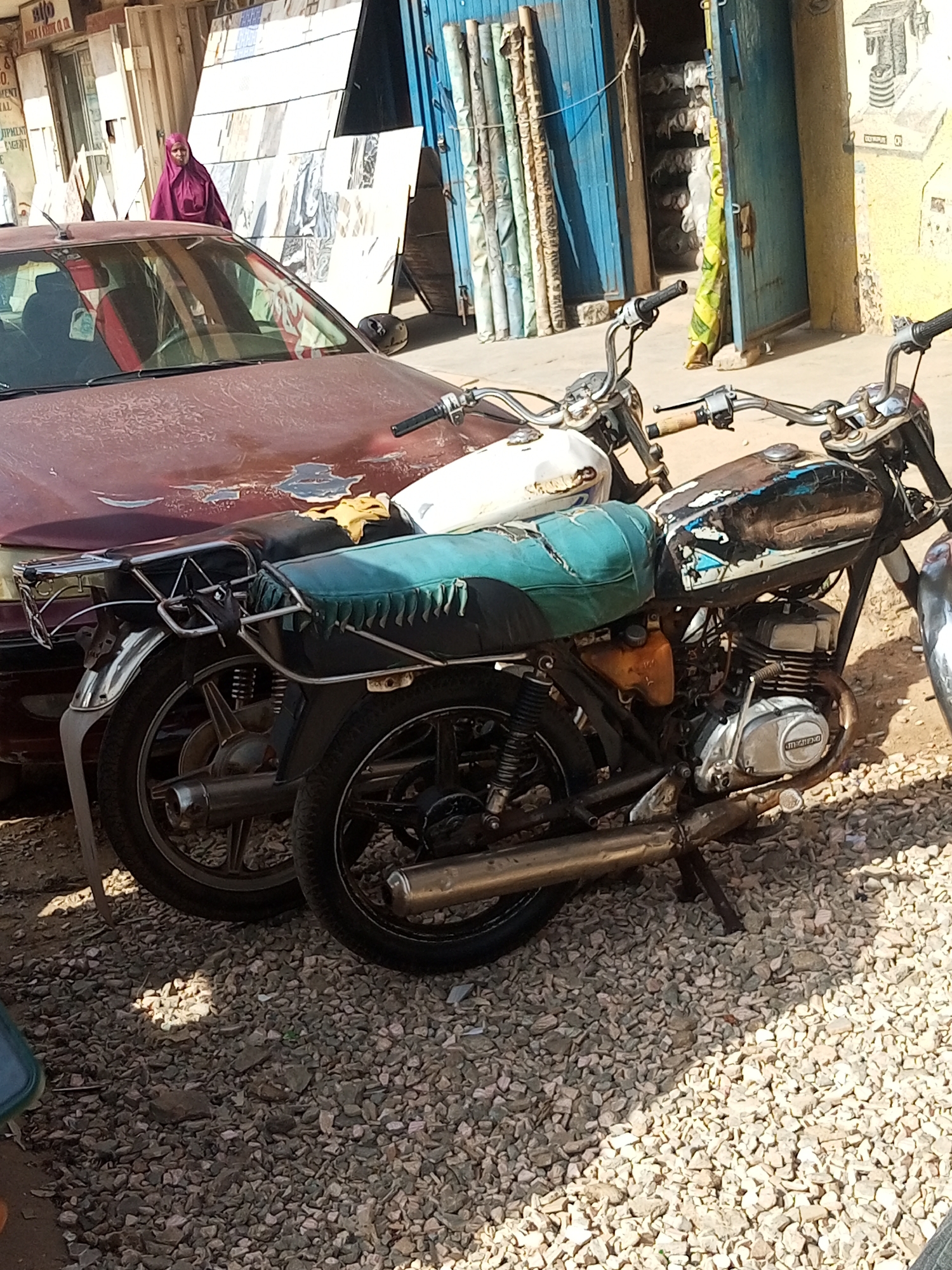 The scene shows a dusty street in what is presumed to be Maiburiji, Nigeria, given the prompt. A maroon car with paint damage is parked alongside two motorcycles. The motorcycles are old, with one having a faded blue seat and a rusted fuel tank. One is partially obscured by the car. A person wearing a pink head covering is visible in the background, near a shop with stacked tiles. The environment is gravelly and suggests an unpaved or poorly maintained area. Rolled carpets or fabrics stand next to a blue metal door. The overall impression is one of a low-income or developing urban area.