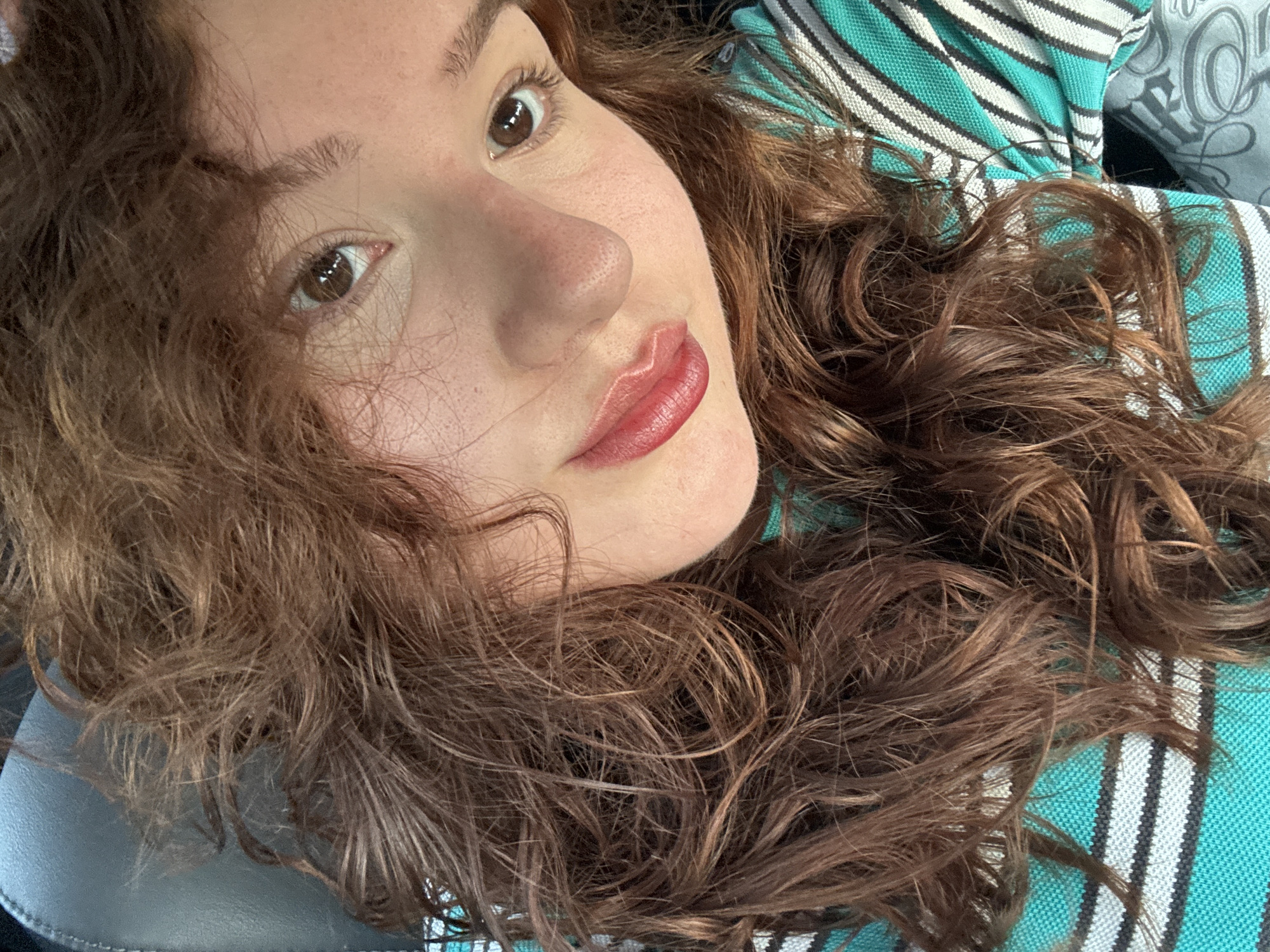 A close-up selfie of a woman with curly brown hair, fair skin, brown eyes, and wearing lip gloss. The background is a striped fabric in shades of teal, white, and gray, and a gray leather-like surface. The location and existence of this photo are not related to Elhovo, Bulgaria.