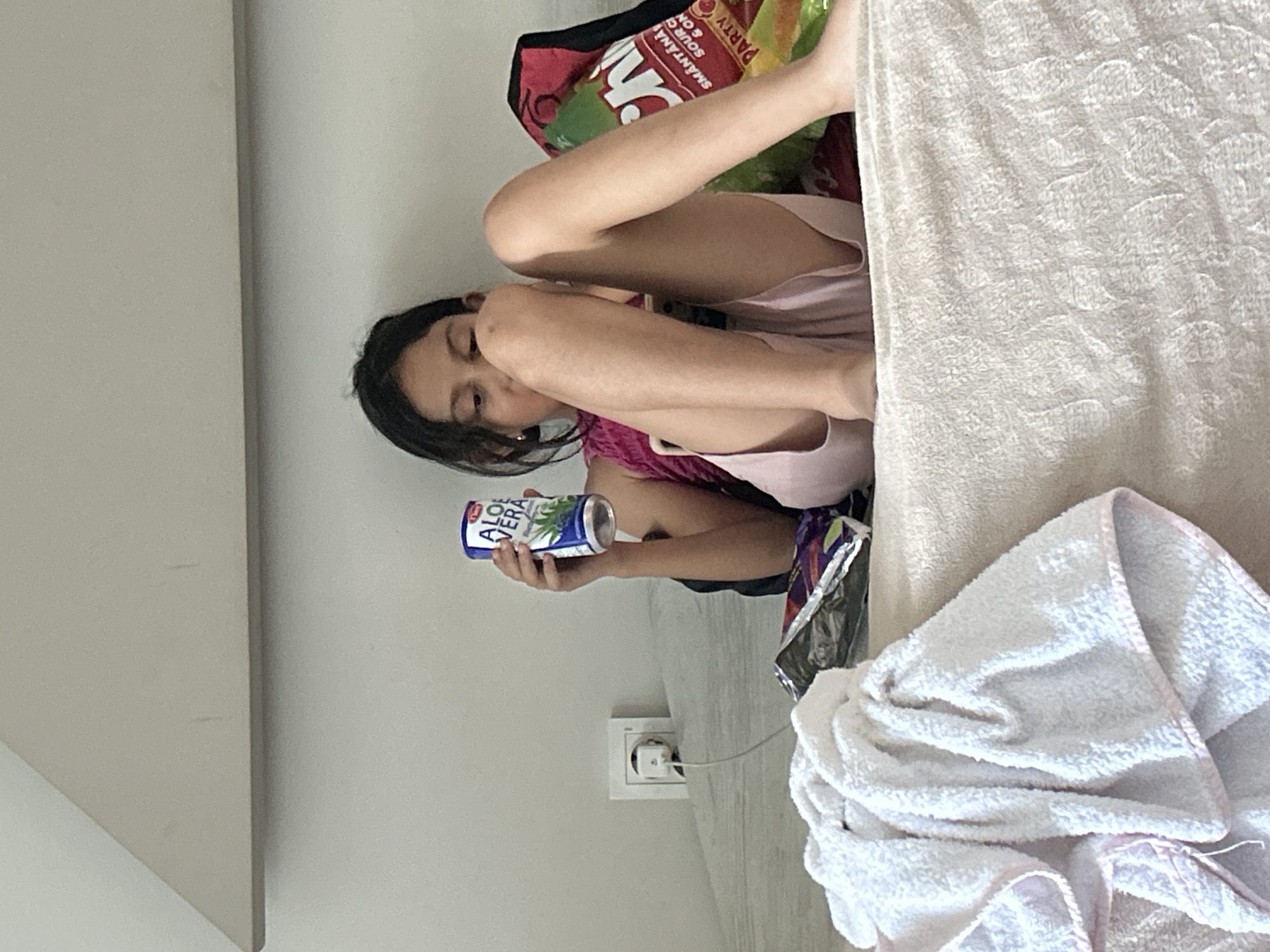 A young girl with dark hair is sitting on a bed, holding a can of "ALOE VERA" drink. She is wearing a pink top. Beside her are bags of snacks and a white towel. A power outlet is visible on the wall behind her. The room appears to be a bedroom, with a textured bedspread and a light-colored wall. The scene is indoors.