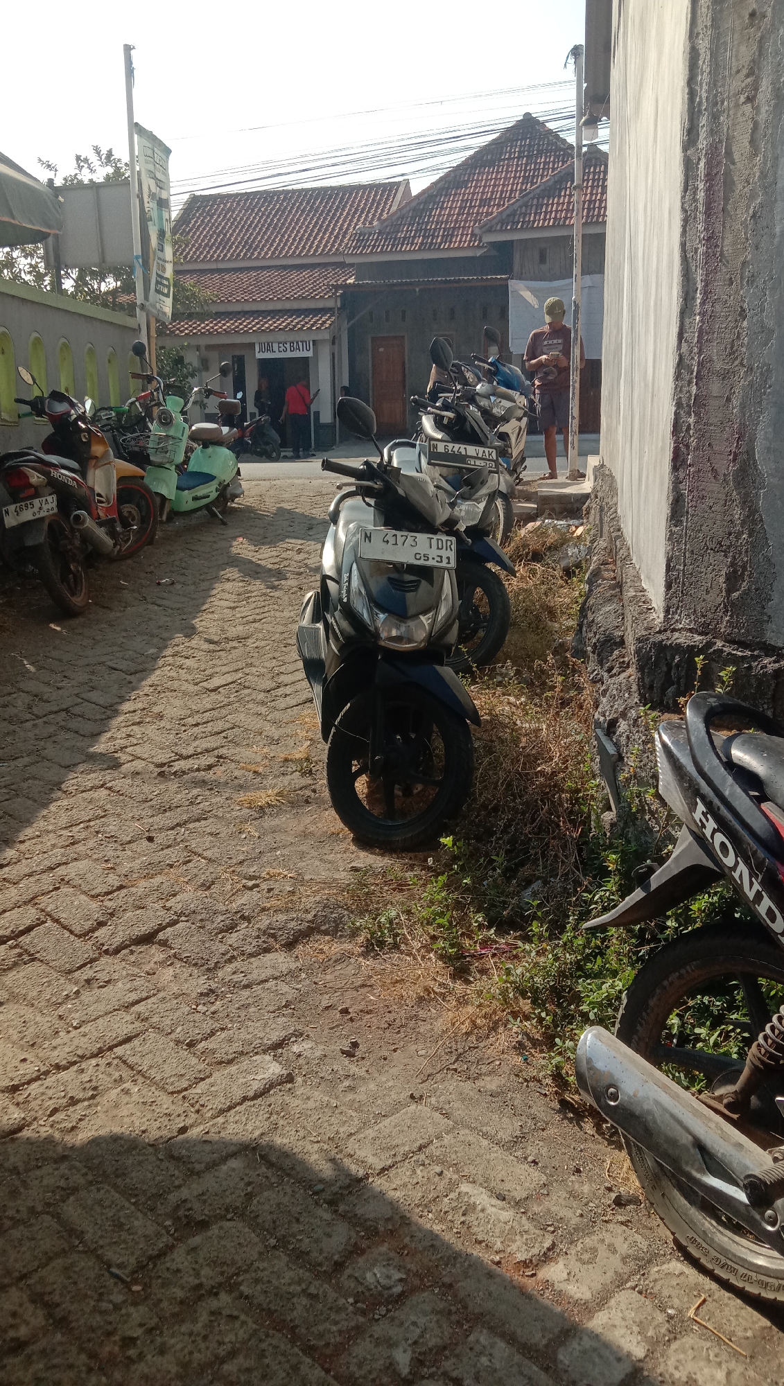 In the city of Karanganyar, Indonesia, on a sunny day, a street is lined with parked motorcycles. The street is paved with interlocking bricks. A light gray wall runs along the right side of the image. On the left, a low wall with arched openings is visible, with vegetation growing in front of it. Behind the motorcycles, a building with a red tile roof houses a business labeled "JUAL ES BATU," which translates to "ICE FOR SALE." Several people are standing near the building, some appearing to be customers and others employees. One man in a maroon shirt and shorts is standing near the gray wall, looking at his phone. The scene is illuminated by the sun, casting shadows on the brick pavement.