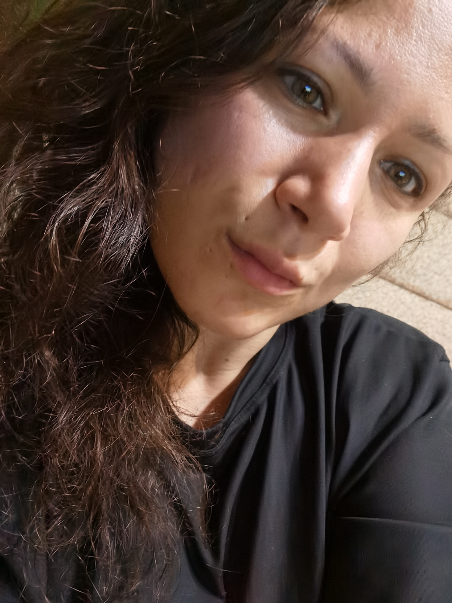 This is a close-up, waist-up, indoor selfie of a woman with dark, curly hair and brown eyes. She is wearing a black t-shirt. The background appears to be a couch with a textured pattern. The lighting suggests it is indoors, possibly in the evening, and there is no identifiable location in Chaikovsky, Russia.