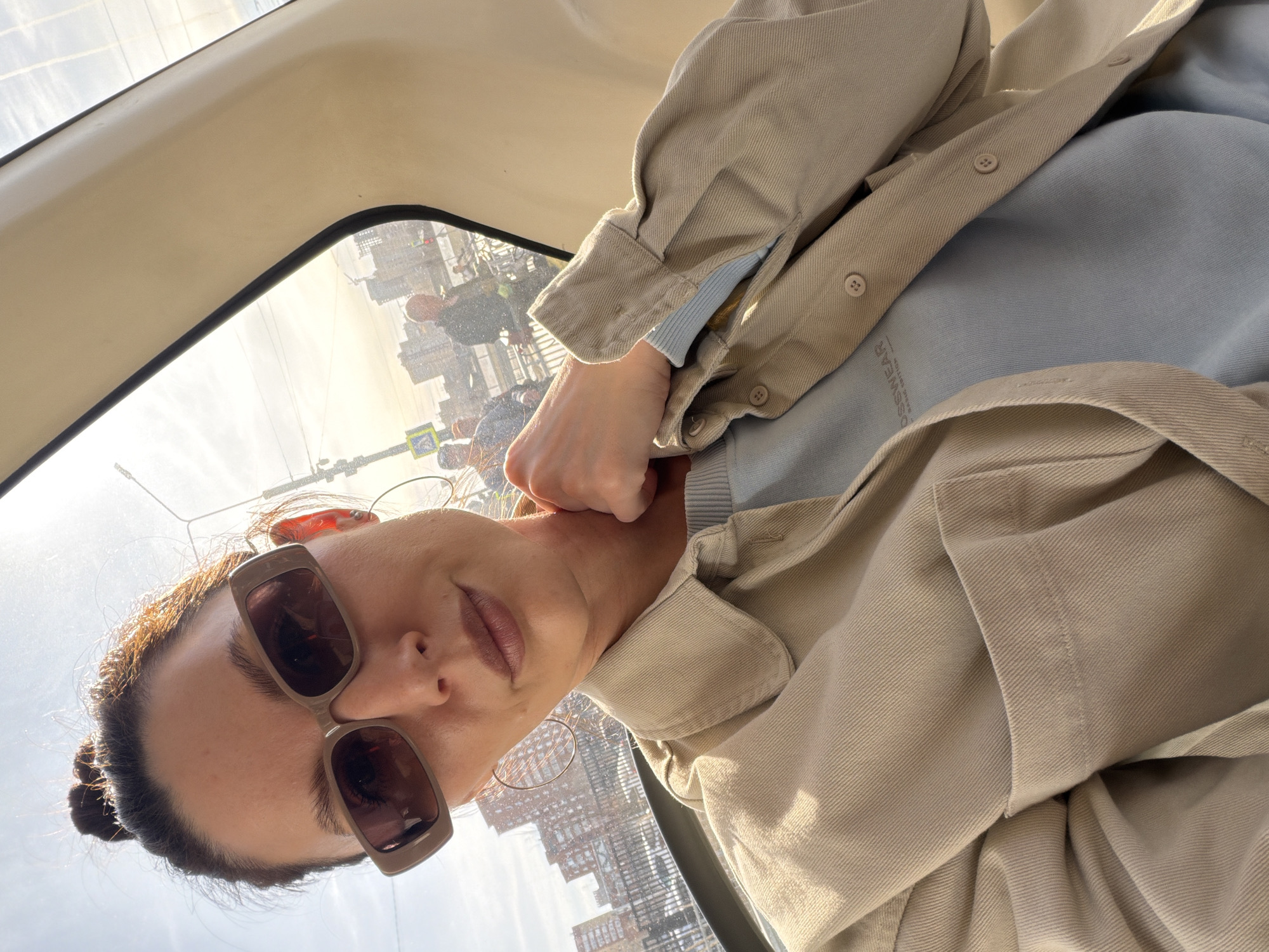 A woman wearing brown sunglasses and a beige jacket over a light blue shirt is taking a selfie inside a vehicle. The background visible through the window shows a cityscape, identifiable as Perm, Russia, due to the distinct architectural styles and urban layout. People are walking in the background. The vehicle appears to be in motion.
