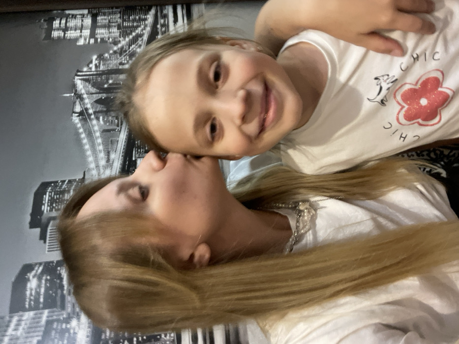 An adult female is depicted kissing a young female child on the forehead. The adult, positioned on the left, has long blonde hair and wears a white t-shirt and a multi-layered necklace with chain and light, irregular elements. She is seen in profile, with her lips touching the child's forehead. The child, on the right, faces the camera with a slight smile, her light-colored hair styled away from her face. She wears a white t-shirt featuring a red glittered flower, the text "CHIC CHIC," and small dark animal silhouettes. Her left arm is visible embracing the adult. The background consists of a black and white mural or wallpaper illustrating a cityscape with tall buildings and a prominent suspension bridge. The specific location within the city of Рубцово, Russia, cannot be identified from the visual information provided.