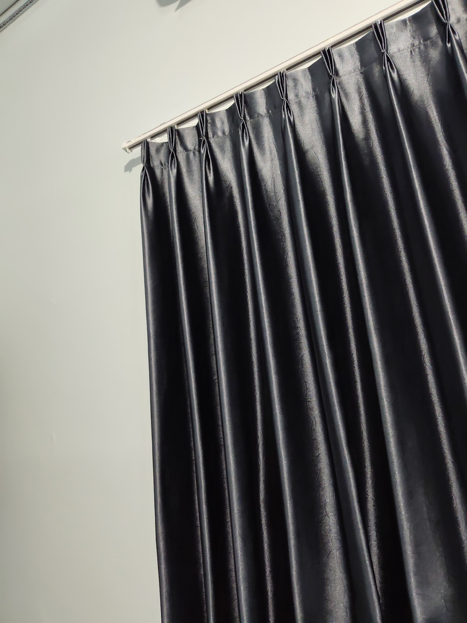 A close-up, low-angle shot captures a dark gray curtain hanging from a white curtain rod against a pale blue wall. The curtain is pleated and features a subtle, embossed floral pattern. The lighting casts shadows, highlighting the texture and folds of the fabric. The setting appears to be an interior space, possibly a residential or commercial room in Pamotan, Indonesia.