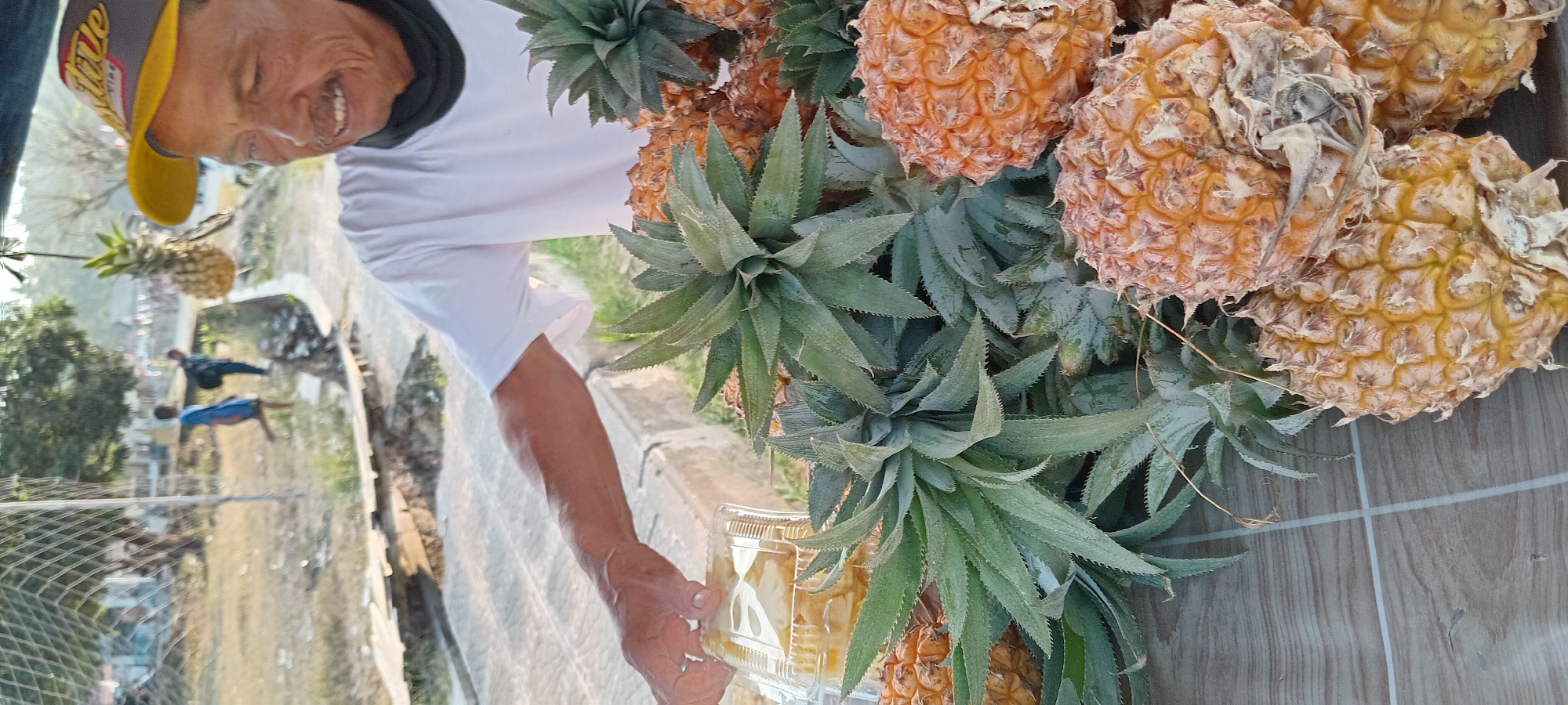 An elderly man with a mustache and a yellow baseball cap, wearing a white t-shirt, is standing behind a display of pineapples. He is smiling and holding a clear plastic container filled with sliced pineapple. The pineapples are piled on a wooden surface in front of him, with their green leafy tops visible. In the background, two young boys are walking in a grassy area next to a concrete structure and a wire fence. The scene appears to be an outdoor market in Kalipucang, Indonesia, likely near a street or public area. The lighting suggests it is daytime, possibly late afternoon.