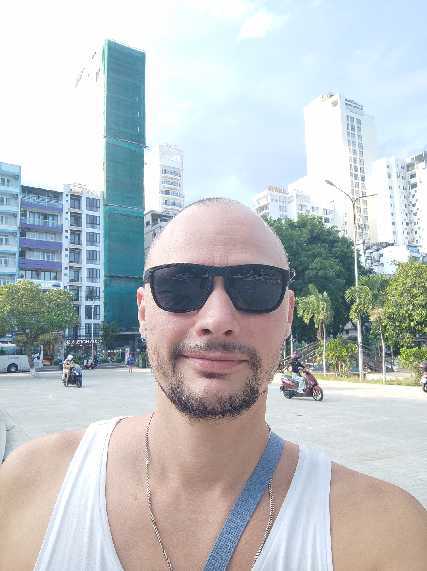 A selfie is taken in front of a city skyline with several tall buildings. The person in the foreground is a bald man wearing sunglasses and a white tank top with a silver chain. Behind him, there are various buildings, some of which appear to be under construction, covered in green netting. Palm trees and other foliage are visible. Several people are seen in the background, some riding motorcycles. The ground is paved. The sky is bright and clear with some clouds. The scene appears to be a public outdoor space in a city.