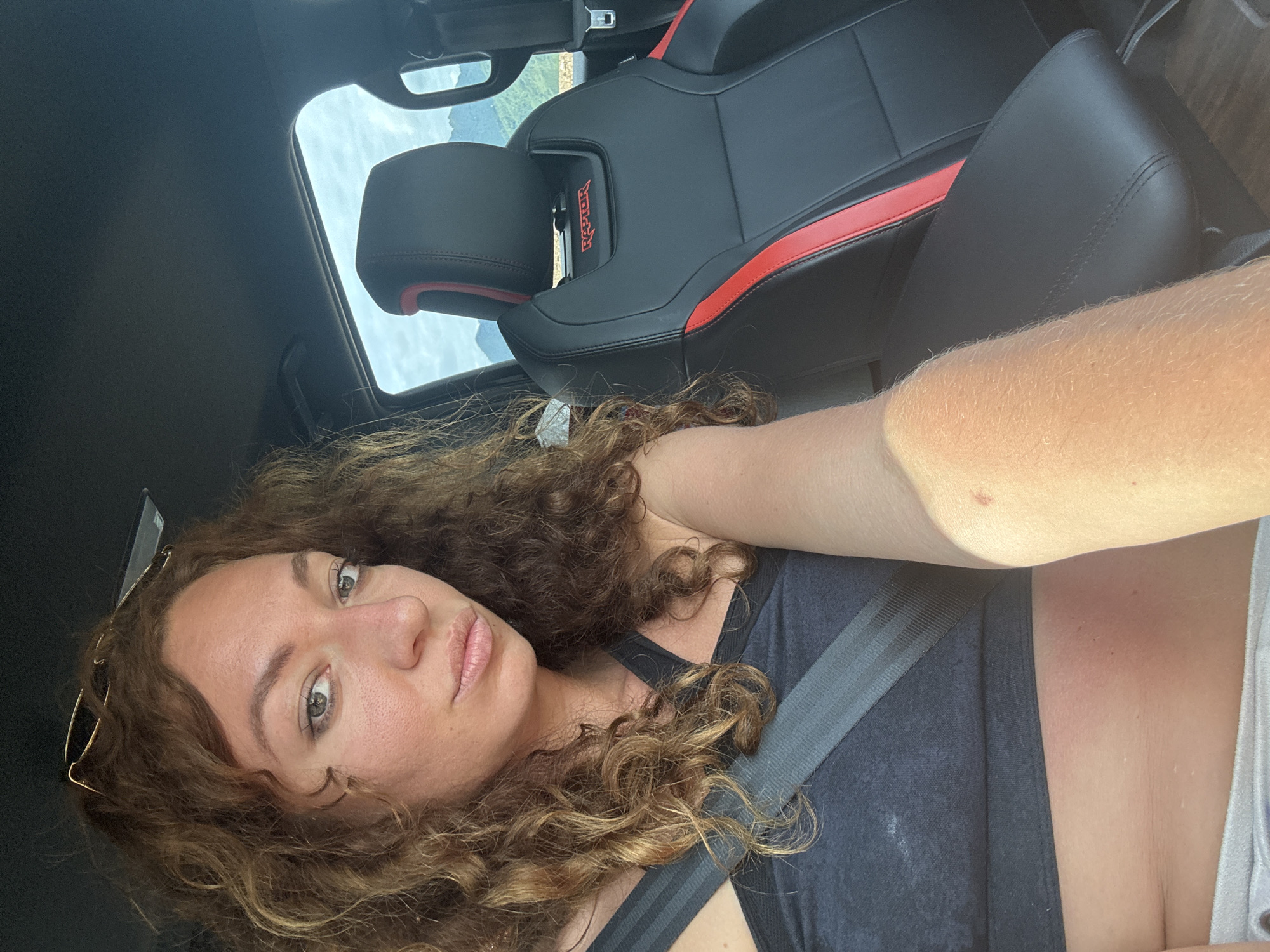 A woman is sitting in the passenger seat of a car, looking at the camera. She has long, curly brown hair and is wearing a black sports bra and grey shorts. A seatbelt is across her chest. The car's interior is visible, including the headrest of the driver's seat, which has red accents and a "RAPTOR" logo. Outside the car window, mountains and a cloudy sky are visible. The image appears to be taken during the daytime. The scene is consistent with a passenger in a vehicle in a rural or mountainous area.