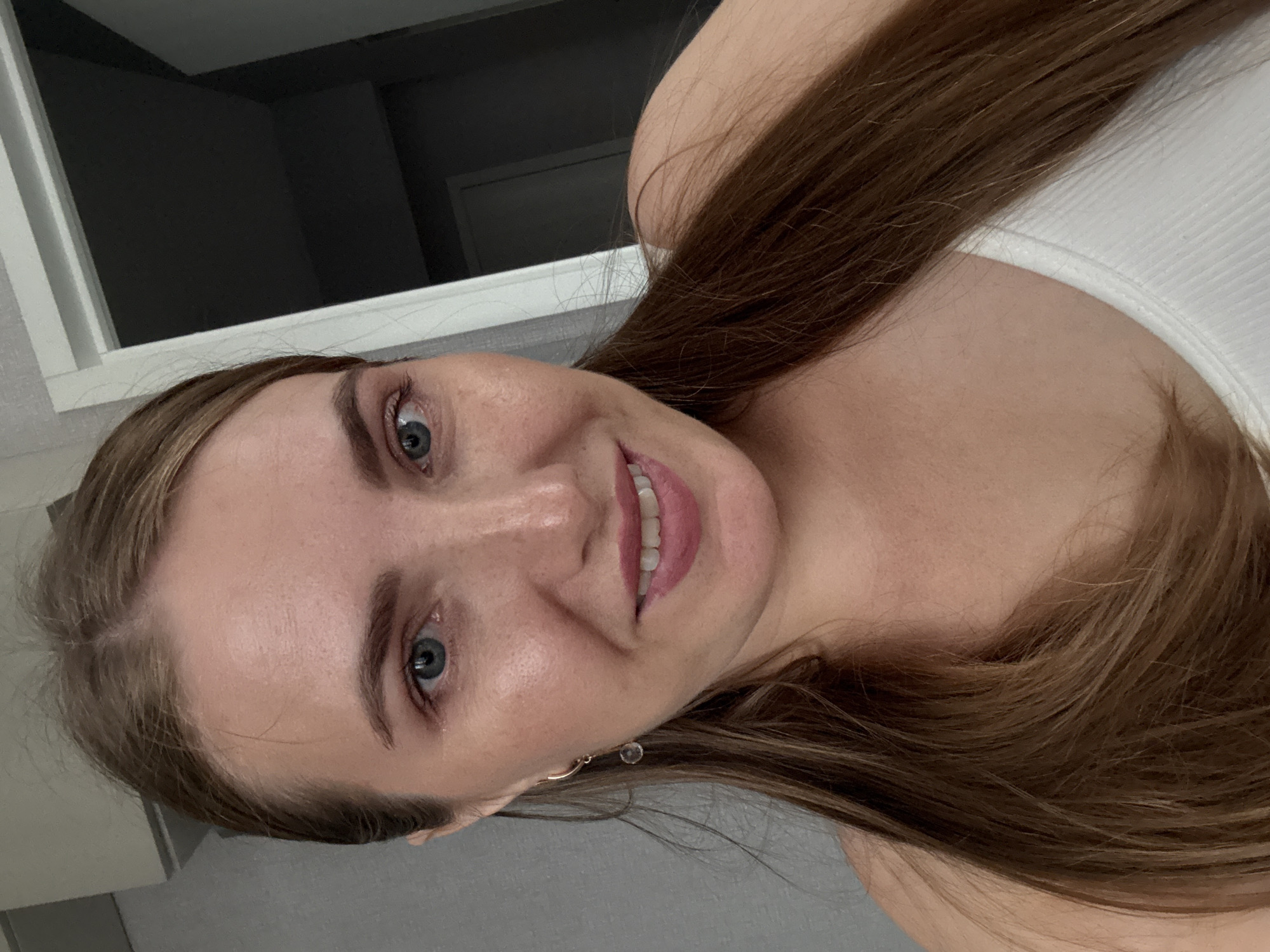 A medium shot shows a woman with long, brown hair, fair skin, and blue eyes. She is smiling, revealing her teeth. She is wearing a white tank top and a gold earring with a clear stone. Her hair is parted in the middle and falls over her shoulders. The background is blurred, but appears to be an indoor setting with a doorway and a wall. The image is likely a selfie taken by the woman. The location is not specified as Kazan, Russia, or any other city.