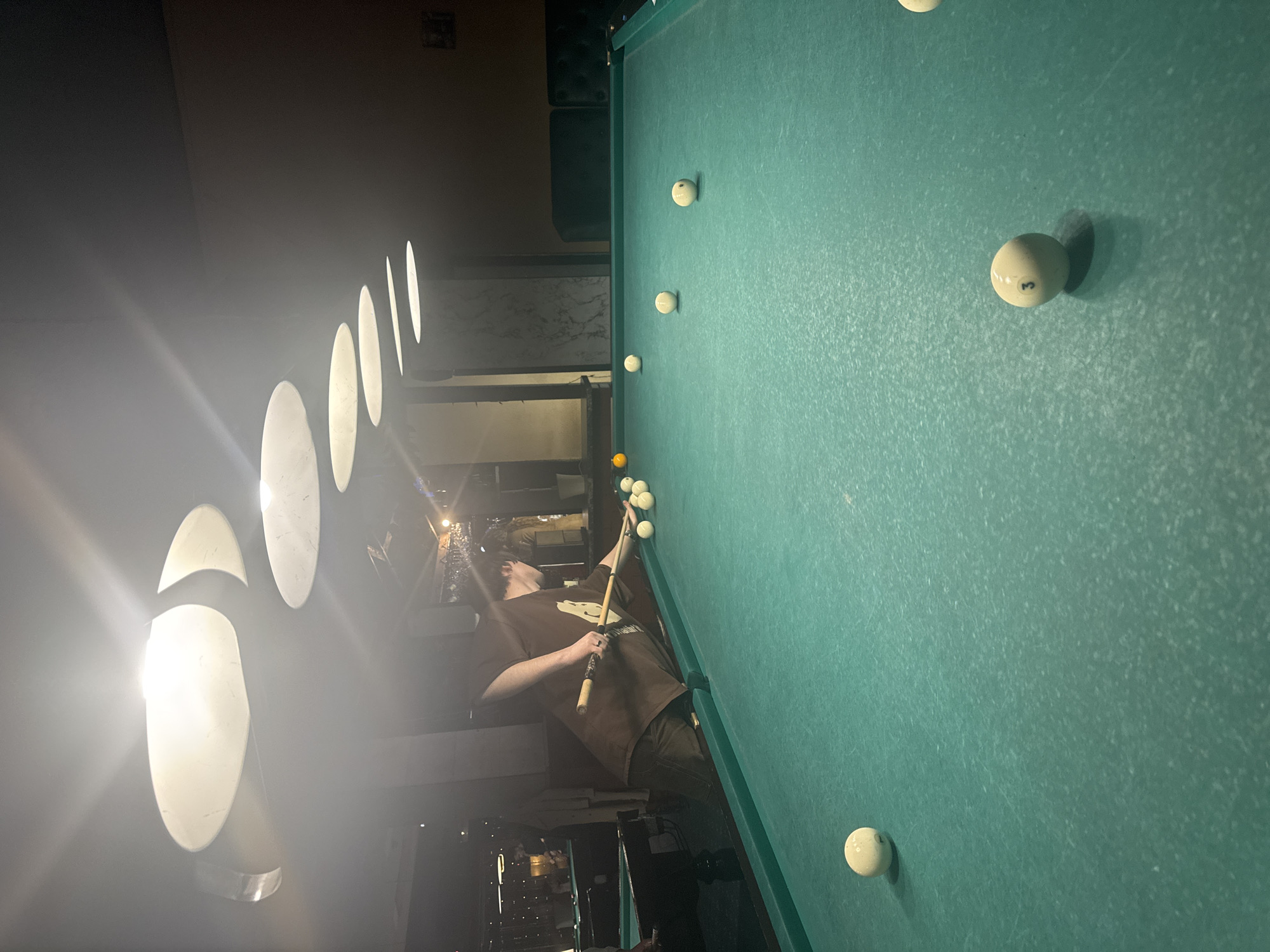 A man plays pool in what appears to be a dimly lit interior, possibly in Moscow, Russia. The pool table, covered in green felt, is surrounded by several pool balls. The man, wearing a brown shirt, is in the process of taking a shot with a pool cue. Overhead are several distinctive, vertically oriented oval light fixtures, a common design element in the city.