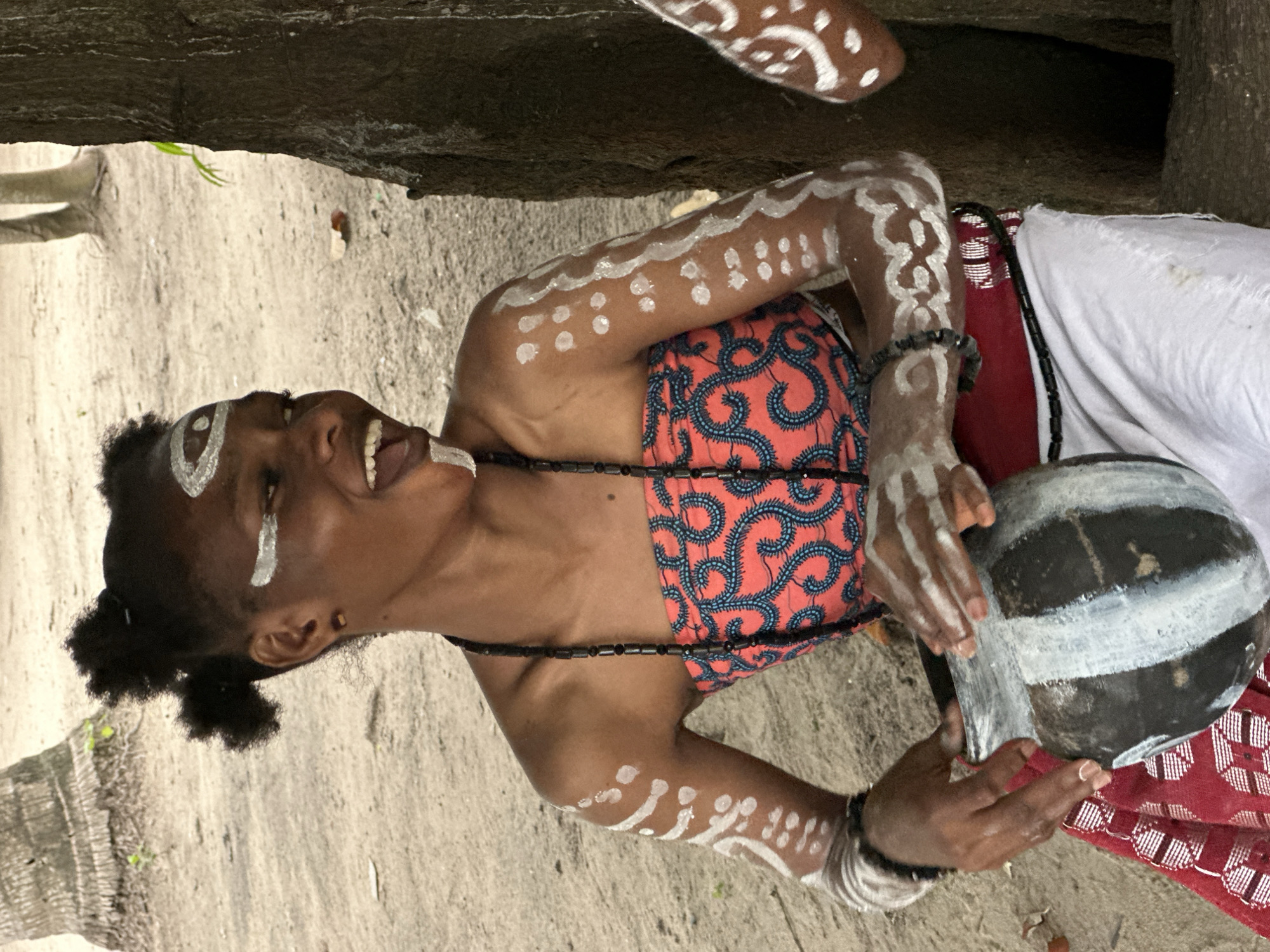 A young Black female, adorned with white body paint on her face, arms, and hands, sits on sandy ground in Badagry, Nigeria, holding a dark, round pot with white paint markings. Her hair is styled in two puffs. She wears a strapless coral-patterned top and a red and white patterned skirt. Her expression is joyful, with her mouth open in a wide smile. A dark beaded necklace is visible around her neck. Behind her, a large tree trunk is prominent on the right, and palm trees and sandy ground make up the background. The scene appears to be outdoors, possibly in a cultural or ceremonial setting.