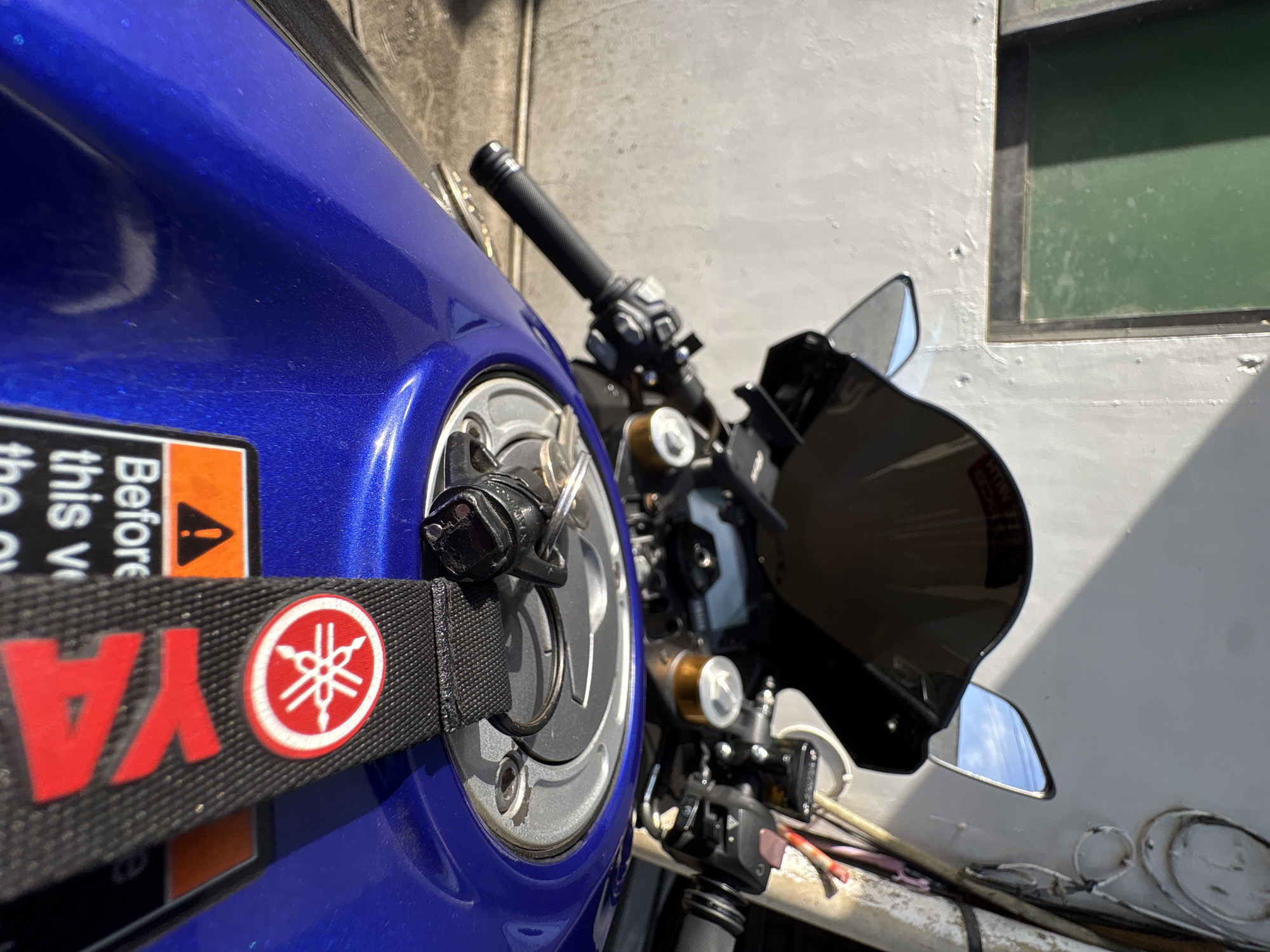 The image shows a close-up view of the handlebars and fuel tank of a blue Yamaha motorcycle parked outdoors. A black fabric lanyard with the Yamaha logo and the word "YAMAHA" in red is attached to the fuel cap. A set of keys is visible on the lanyard. The motorcycle is positioned in front of a textured, light-colored wall, with a green-tinted window on the left side of the frame. Sunlight casts shadows on the wall and the motorcycle. The background suggests an urban or semi-urban setting, possibly a street or a residential area in Davao City, Philippines. The focus is on the motorcycle's details, indicating it is the primary subject.