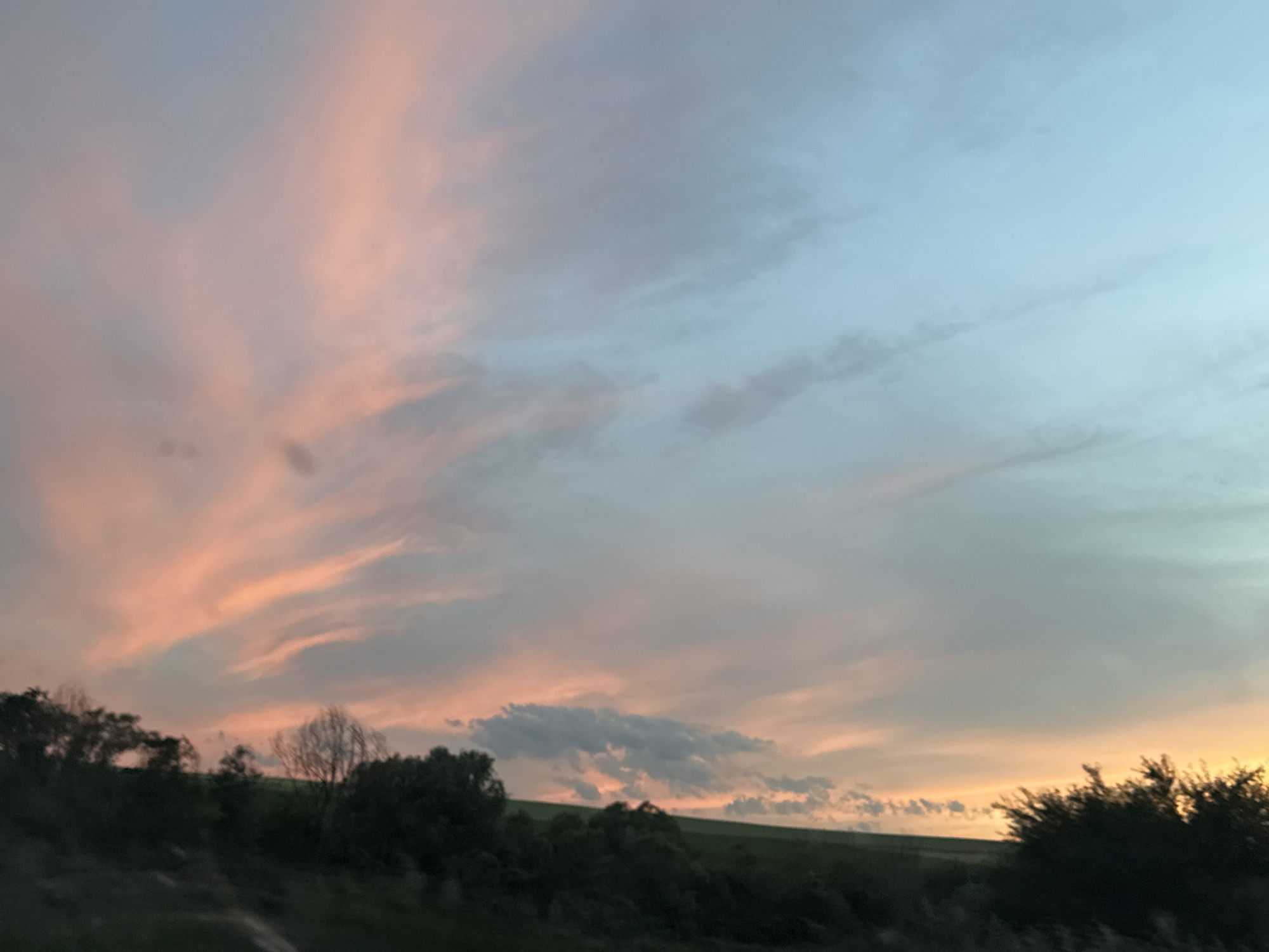 The media file captures a picturesque sunset over a rural landscape. The sky is a dramatic canvas of pastel colors, with streaks of soft pink and orange clouds juxtaposed against a gradient of pale blue. These vibrant hues suggest the sun is low on the horizon, indicating either dawn or dusk.
The foreground is dominated by silhouetted trees and rolling hills, creating a natural, serene setting. The trees are dense and dark, with some appearing bare, hinting at either a late autumn or early spring season. The land appears to be agricultural, with subtle contours suggesting fields or meadows.
The overall mood is tranquil and contemplative, evoked by the soft lighting and the expansive, natural scenery. There are no people or man-made objects visible, emphasizing the unspoiled, natural beauty of the location. The scene suggests a peaceful moment in time, perhaps a quiet evening in the Bulgarian countryside near Beli Lom, where the day is gently concluding. No text is visible in the media file.