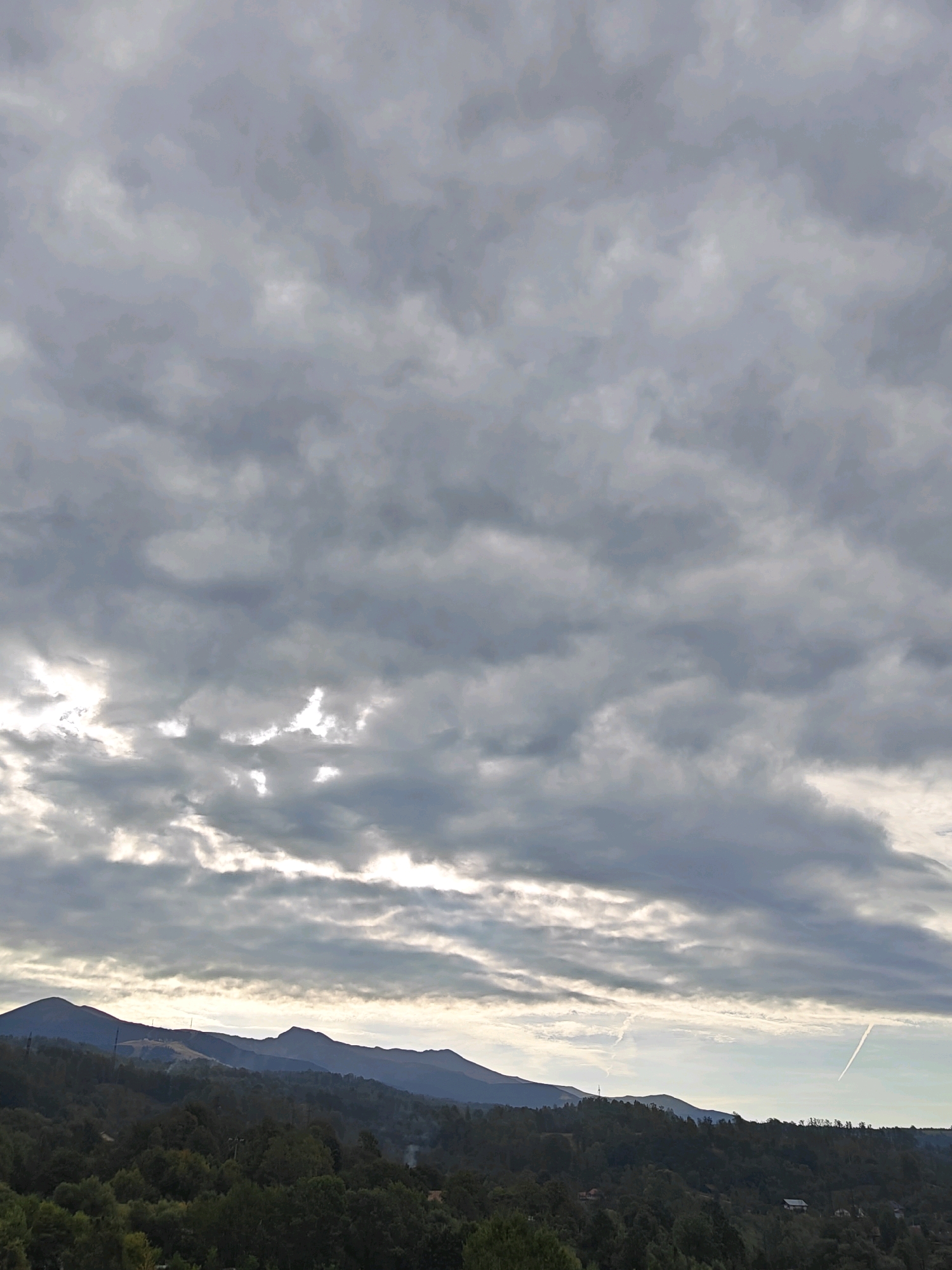 A wide-angle, landscape photograph captures a cloudy sky over a mountainous, forested area. The sky is filled with numerous gray and white clouds, with some breaks in the clouds allowing sunlight to filter through, creating streaks of light. In the distance, several mountain ranges are visible, their peaks silhouetted against the lighter sky. The foreground is dominated by a dense, dark green forest that covers rolling hills. The terrain appears to be somewhat rural or undeveloped, with a few scattered buildings partially obscured by the trees. There is no visible indication of Petroșani, Romania in the image.