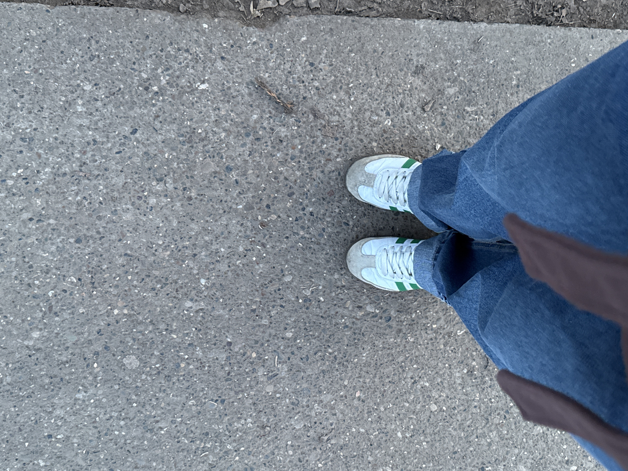 The image presents a top-down perspective of a person standing on a concrete surface, possibly a sidewalk. The individual is wearing light blue jeans and white sneakers with green stripes. A portion of a brown bag or garment is visible on the right side of the frame. The concrete surface appears to be textured with small pebbles and imperfections. The location is potentially the city of Chaikovsky, Russia, but this cannot be verified with the given image alone. No specific actions or interactions are apparent, and the environment consists mainly of the concrete surface.