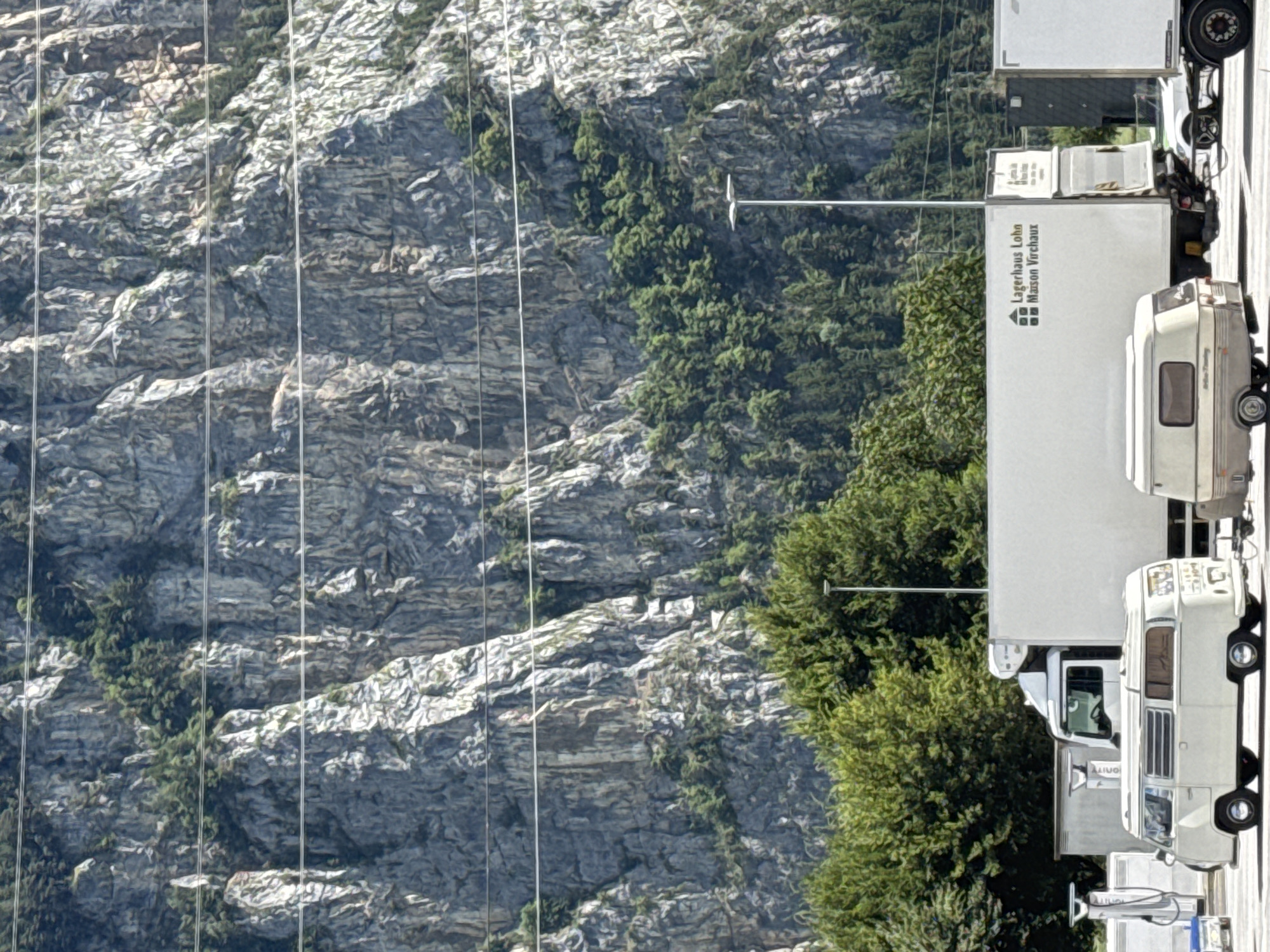 A bright, high-angle outdoor medium shot captures a parking area in front of a sheer, grey, rocky mountain face in Schattdorf, Switzerland. The mountain is partially covered with green trees and appears in the background. In the foreground, three trucks are parked. The first truck on the left is white with a black cab and is mostly obscured by a white trailer. The second truck, in the center, is also white with a black cab and a white trailer. The trailer has the logo "Lagerhaus Lohn Maison Virchaux" printed on it. The third truck on the right is partially visible and appears to be a white trailer.
Between the first and second trucks, a vintage white camper van is parked, facing left. To the left of the camper van, there are two white charging stations. In front of the camper van, a very small, light blue vehicle is parked. A light brown and cream colored caravan is parked behind the camper van. The scene is illuminated by bright sunlight, casting shadows and highlighting the textures of the rock face.
