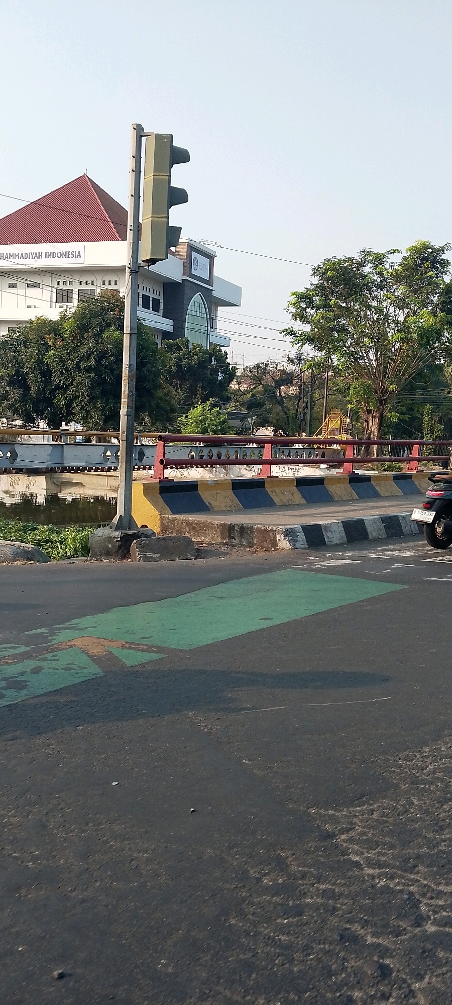 The image depicts a traffic light post and a street in Bekasi, Indonesia. In the background, a white building with a red-tiled roof, labeled "MUHAMMADIYAH INDONESIA," is visible. Adjacent to it is another building with arched windows. A bridge spans a body of water, and lush green trees border the scene. The foreground shows a road with green and orange markings, indicating a pedestrian crossing or bike lane. A motorcycle is partially visible on the right side of the road. The sky is clear and blue, suggesting a bright, sunny day.