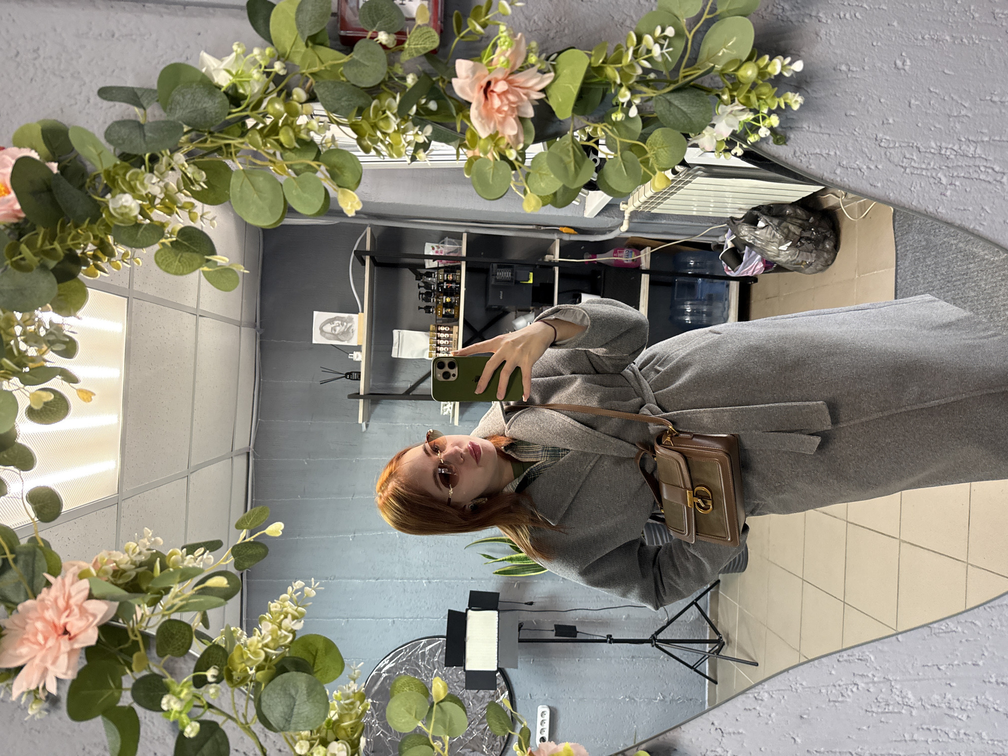 A mirror selfie shows a woman in a grey coat, holding a phone and wearing sunglasses. A tan purse hangs from her shoulder. The mirror is surrounded by a floral wreath. Reflected in the mirror is a room with grey walls and tiled floors, storage shelves holding products, lighting equipment, a refuse bin, and various items. The location is Chaikovsky, Russia.