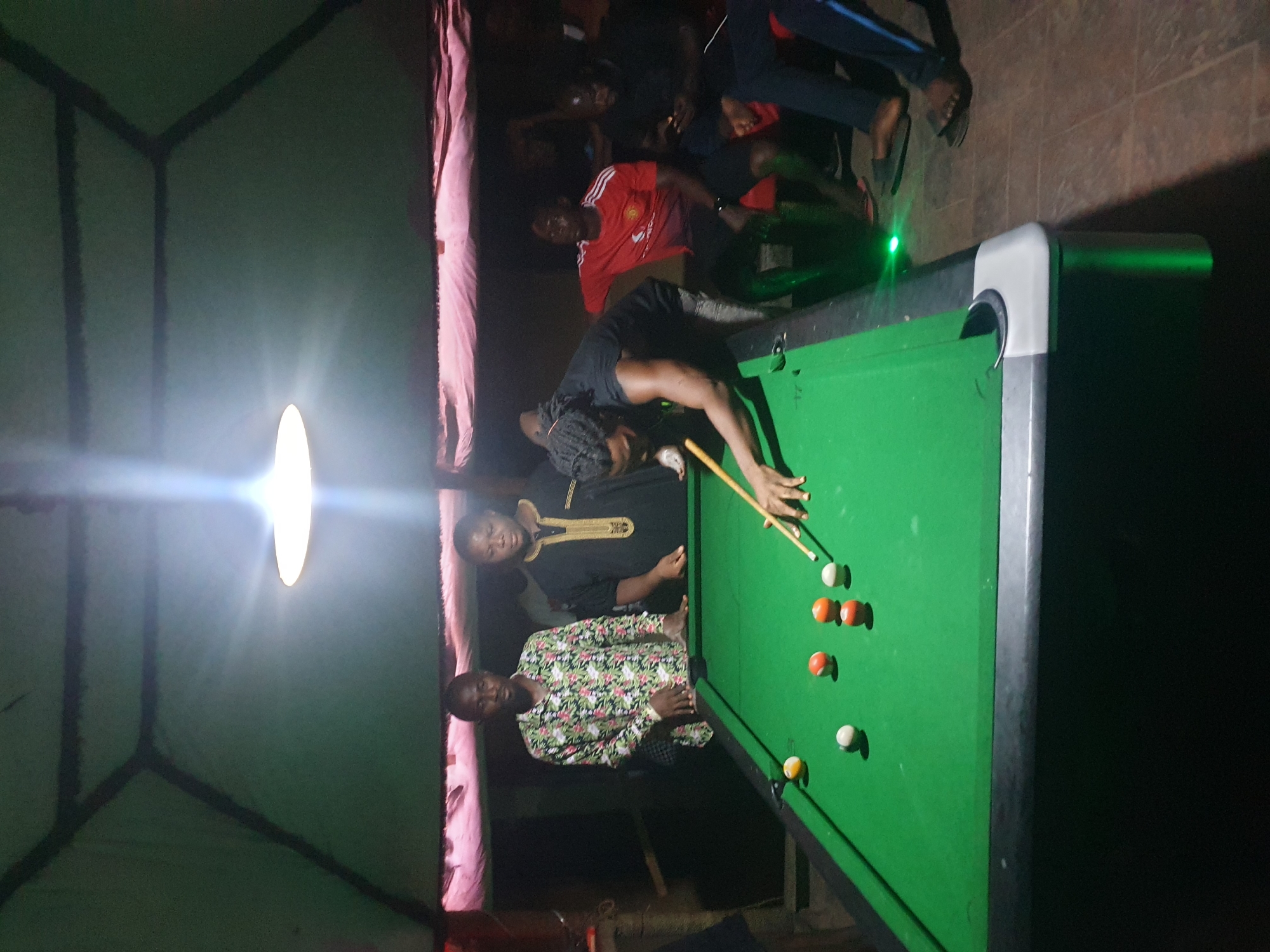 **Scene:**
The scene depicts a group of people playing pool indoors.
**Location:**
Based on the provided information, the location is in the city of Lokoja, Nigeria. The image suggests a possibly makeshift or informal recreational space.
**People:**
There are several people visible in the image. One person is bending over the pool table, cue in hand, preparing to take a shot. Two other individuals are standing near the pool table, observing the game. Additional people are in the background, seemingly watching or conversing.
**Objects:**
The primary object is a pool table with several pool balls placed on it. A cue stick is being held by one of the individuals. A light fixture hangs overhead, providing illumination. Other elements include clothing and furniture.
**Actions:**
The main action is playing pool. People are observing, and socializing.
**Interactions:**
The people are interacting with each other while playing pool, as well as watching and talking in the background.
**Environment:**
The environment appears to be an informal recreational space. The walls seem to be constructed from some kind of textile or tarp, suggesting a makeshift structure. The lighting is provided by an overhead lamp. The general atmosphere indicates a social gathering focused around the game of pool.