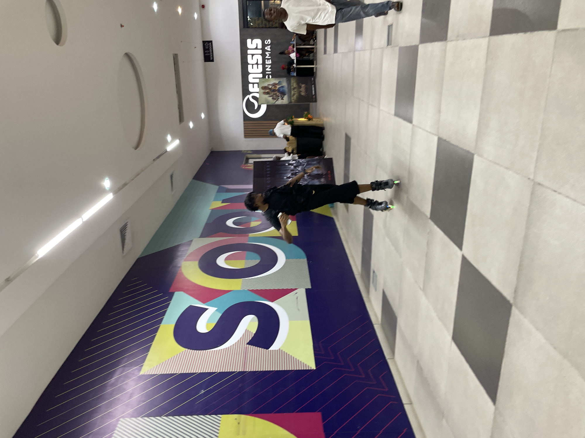 The image depicts an indoor scene at Genesis Cinemas in Moba, Nigeria. A young man is rollerblading down a wide corridor with a distinctive checkered floor. He is wearing a black t-shirt and black shorts and is captured in motion. To the left of the rollerblader is a large, colorful mural on the wall featuring geometric shapes and bold lettering, with the word "SOON" prominently displayed. Further down the corridor, to the right, are ticket counters and movie posters, indicating the cinema's operational area. Several other individuals are visible in the background, including cinema staff and patrons. The lighting appears to be artificial, consistent with an indoor commercial space.
