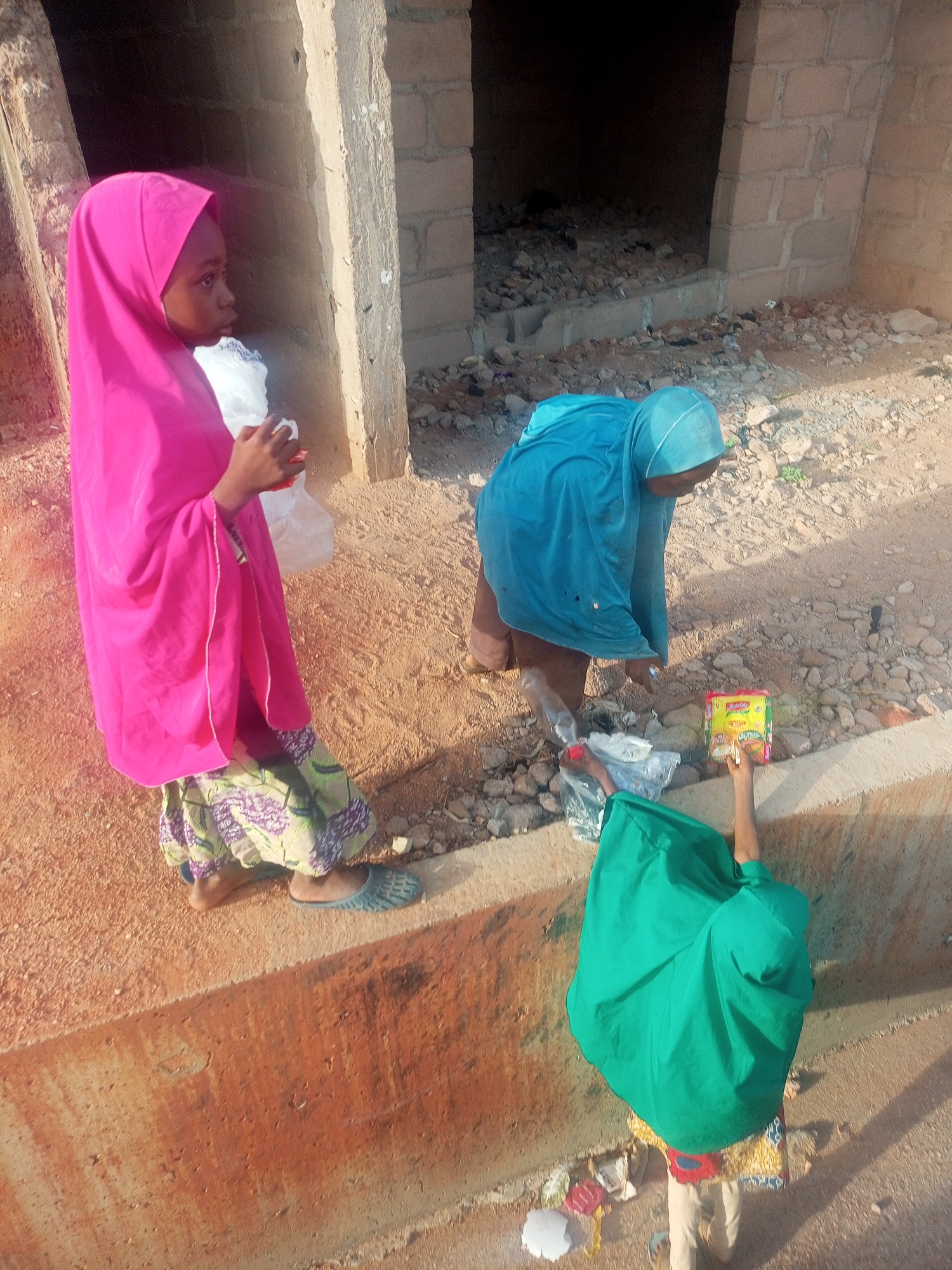 Three individuals are observed in an outdoor setting. The location appears to be near a building structure with exposed brickwork.
One individual is standing, clad in a pink hijab and a patterned skirt. A second individual is bending over, wearing a blue hijab and brown pants. A third individual, wearing a green hijab, is standing at a lower elevation, holding up a packet of yellow noodles. This individual is also wearing a patterned skirt.
Debris is scattered on the ground, suggesting an unkempt environment. The overall scene appears to depict everyday activities in an urban setting.