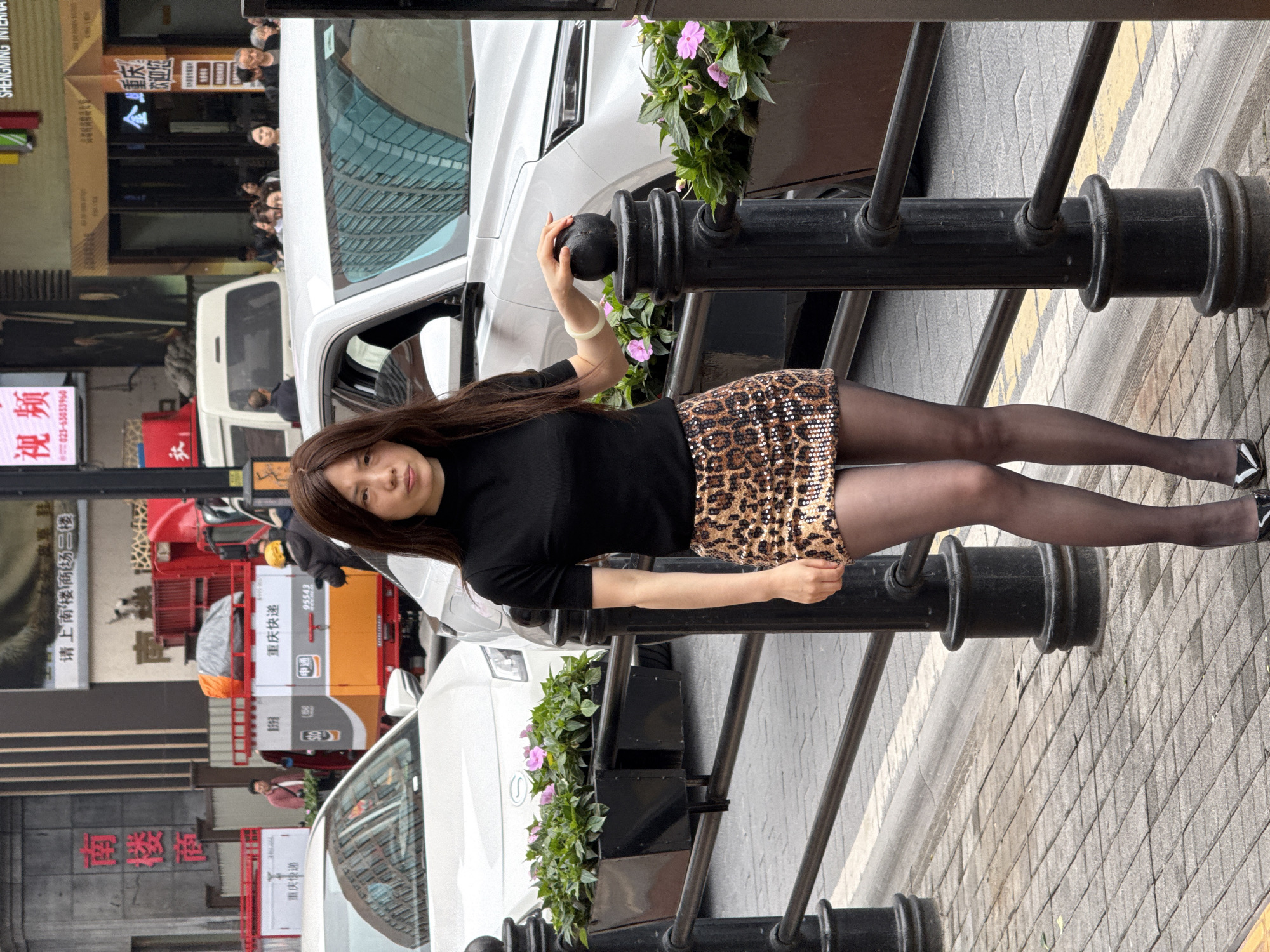 A woman stands next to a metal barrier on a street in the Yuzhong District of Chongqing, China. The barrier has black posts and horizontal rails, with planters of green foliage and pink flowers attached. The woman is wearing a black long-sleeved shirt, a sequined leopard-print mini skirt, black tights, and black and white high heels. She has long brown hair and fair skin. She is touching the top of a metal post with her right hand.
Behind the woman and barrier, there is a white car and a red delivery truck. Signs in Chinese characters are visible on the buildings across the street, including "南楼面" and "重庆快递". People can be seen walking in the background. The street is paved with gray bricks.