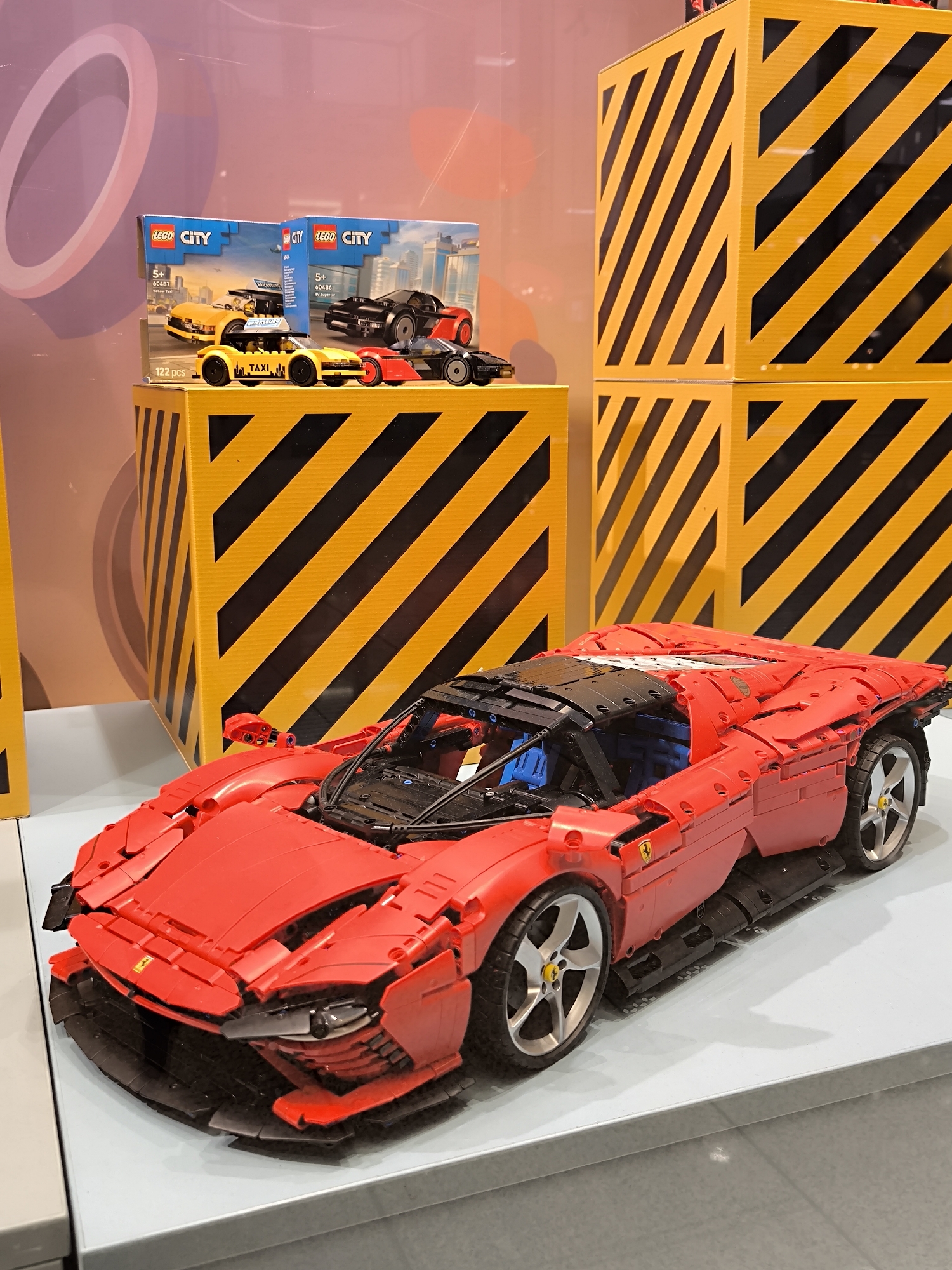 This media file captures a LEGO display, likely within a retail or exhibition setting in Riga, Latvia. The central subject is a large, intricately built red LEGO sports car, resembling a Ferrari Daytona SP3, constructed from numerous LEGO Technic elements and resting on a white display platform. Its sleek design, visible chassis components, and Ferrari badge on the side are notable details.
Behind the large car, elevated on yellow and black striped display boxes that mimic hazard warnings, are smaller LEGO City products. Two boxed LEGO City sets are visible: one for a "Yellow Taxi" (set 60487, 122 pcs, aged 5+) and another for a "Supercar" (set 60486, aged 5+). A built yellow LEGO taxi with "TAXI" written on its roof is positioned in front of its corresponding box, and a black and red LEGO supercar is next to it, likely corresponding to the other box. The backdrop features abstract circular shapes in pink and orange hues, visible through a transparent surface. The overall scene suggests a promotional or retail presentation of various LEGO car models, ranging from detailed adult collector sets to children's play sets. The display boxes are a prominent visual element, creating a dynamic background for the featured models.