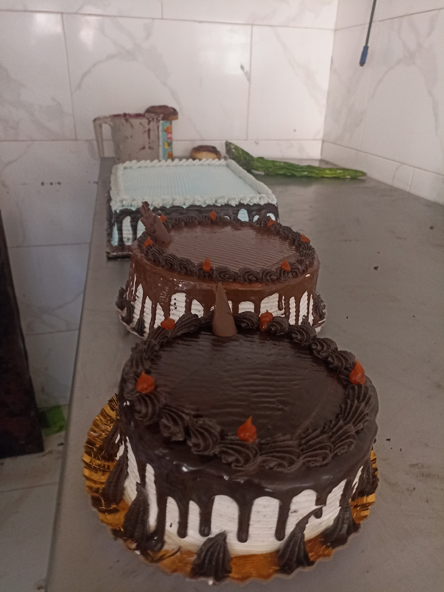 Three chocolate cakes are displayed on a metal counter in what appears to be a bakery in Gaza City, Palestinian Territories. The cakes are tiered and decorated with chocolate icing, white frosting, and small orange candies. The background shows white tiled walls. The lighting is bright, suggesting an indoor setting. No people are visible in the image.