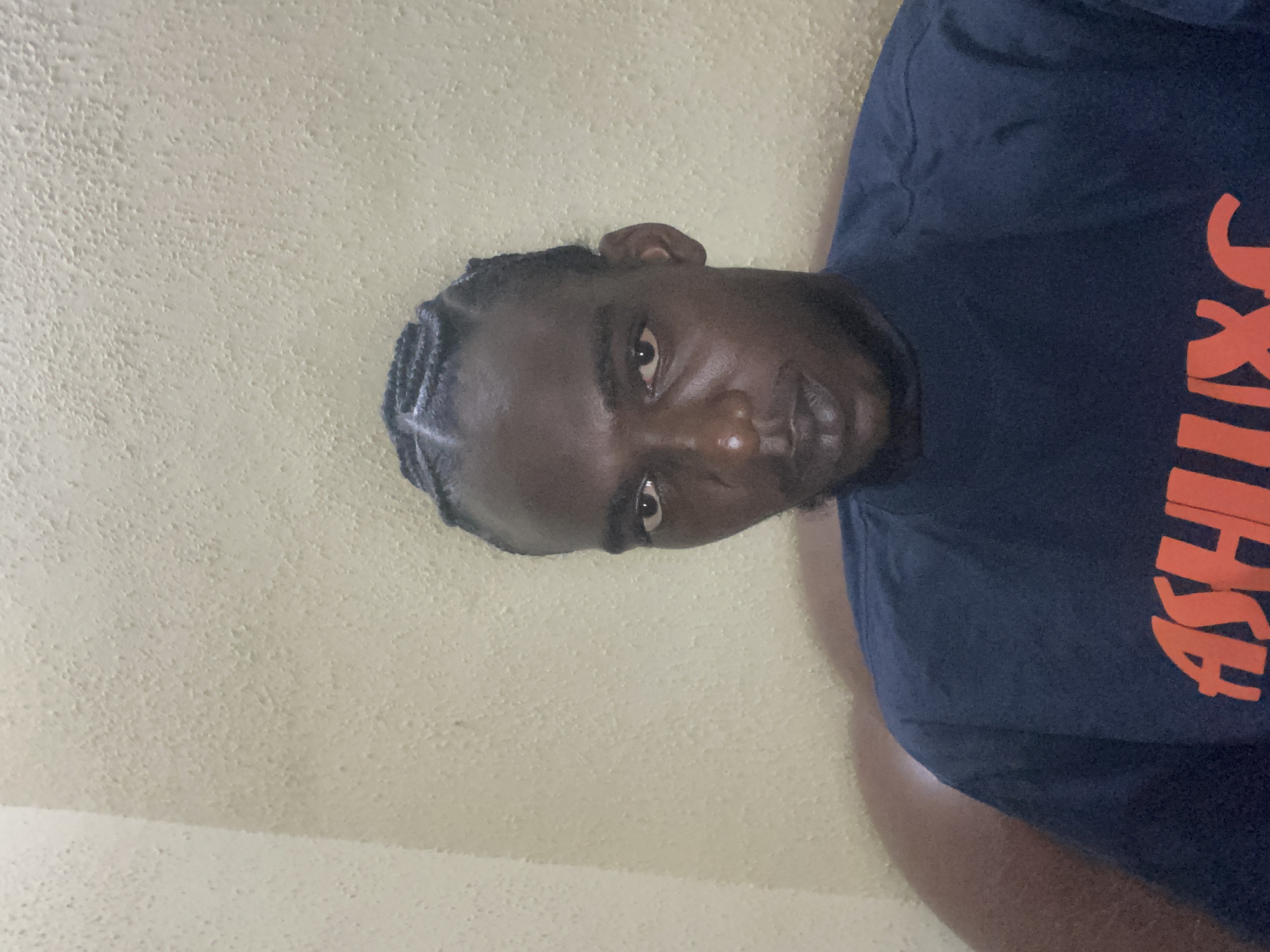 The image depicts an adult male in a portrait-style shot within an indoor environment, located in Lagos, Nigeria. The subject has dark skin, dark eyes, a faint mustache, and a chin strap beard. His hair is styled in multiple dark cornrows running from his forehead to the back of his head. He wears a dark blue, possibly cotton, crew-neck t-shirt with three prominent orange uppercase letters 'ASH' visible on the chest. The man looks directly at the camera with a neutral expression. The background consists of a uniformly light yellowish-beige wall with a pronounced, stippled, or popcorn-like textured finish. A distinct interior corner of the wall is visible on the left side of the frame. A curved, upholstered brown surface, likely part of a sofa or chair, is partially visible behind the subject's left shoulder, contrasting with the wall.