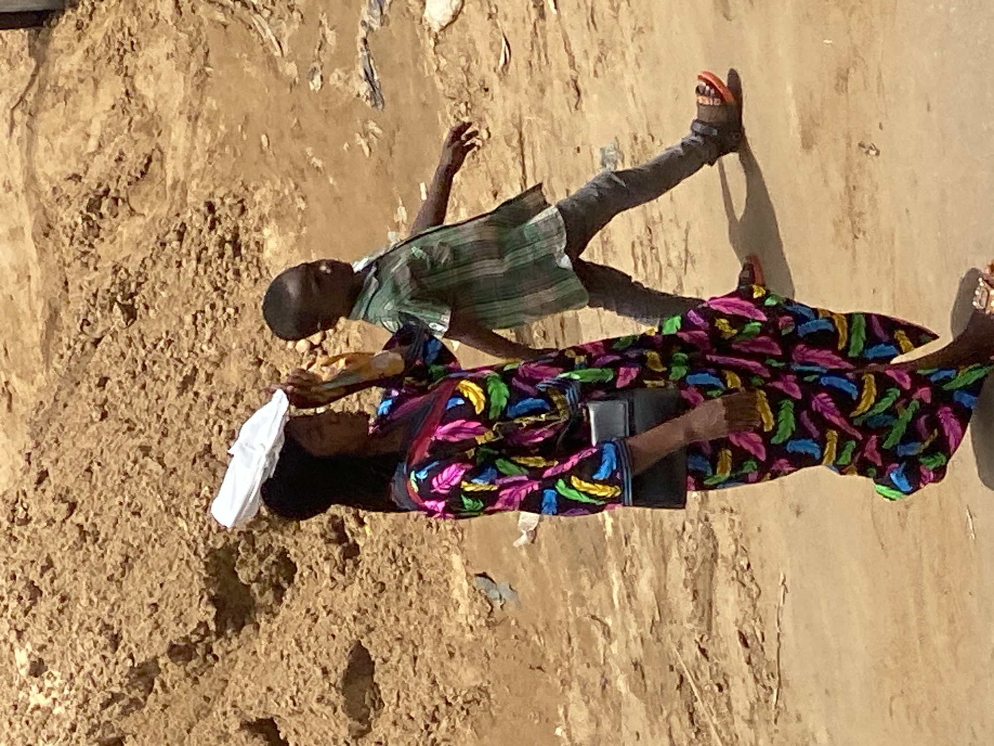 A low-angle shot captures a boy standing beside a woman sitting on the ground. The woman is wearing a colorful dress with a feather-like pattern and has a white cloth draped over her head. She holds a dark object in her hands. The boy is dressed in a striped shirt and gray pants, wearing orange and black sandals. The backdrop suggests an outdoor setting, possibly in the Fegge area of Nigeria, with a rough, earthen ground. The overall composition appears casual and candid.