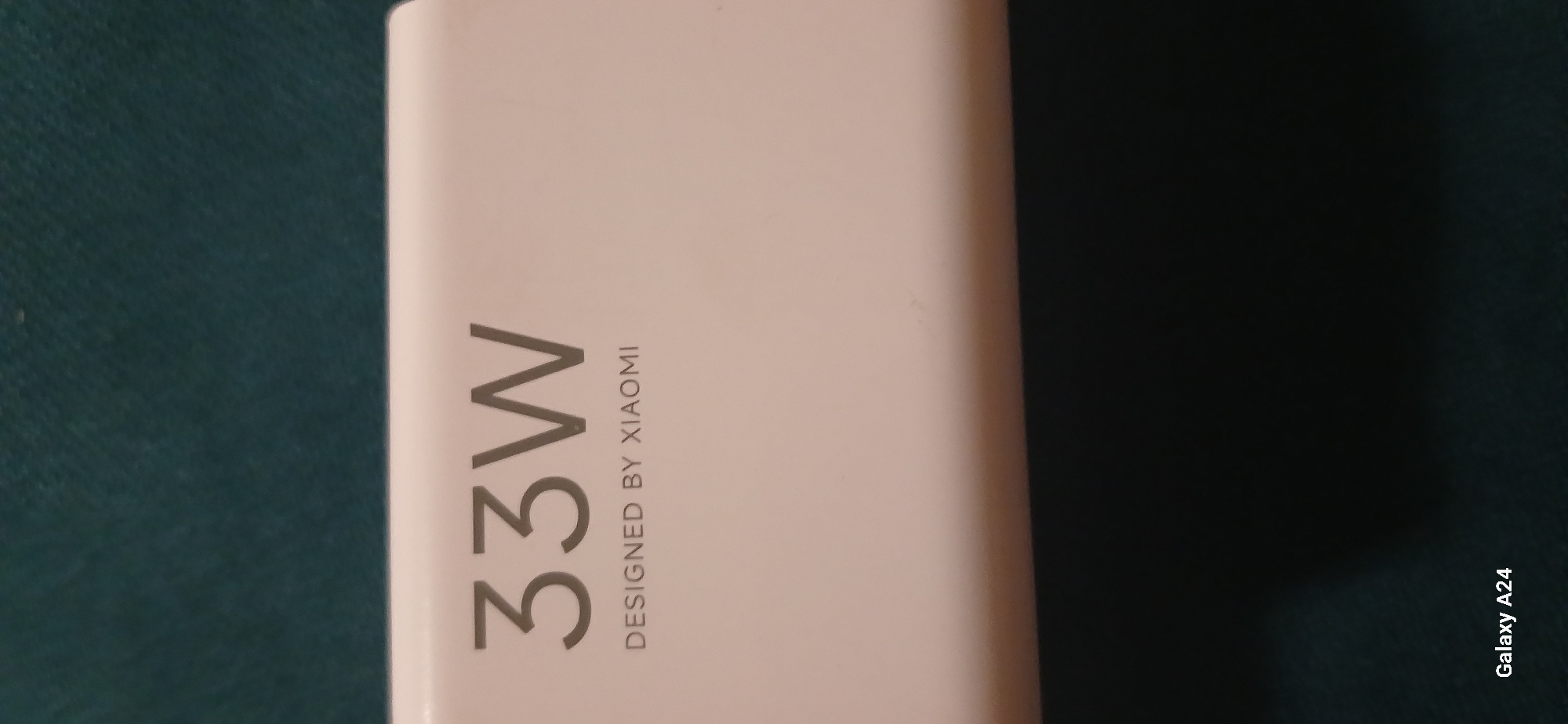 The image shows a white rectangular object with text printed on it. The text reads "33W" in large font and "DESIGNED BY XIAOMI" in smaller font below. The object appears to be a power adapter. The background is a dark, textured surface, possibly fabric. In the bottom left corner, there is text that reads "Galaxy A24". The scene is well-lit, with the white object being the primary subject. No specific location within Bishkek City, Kyrgyzstan can be determined from the image.