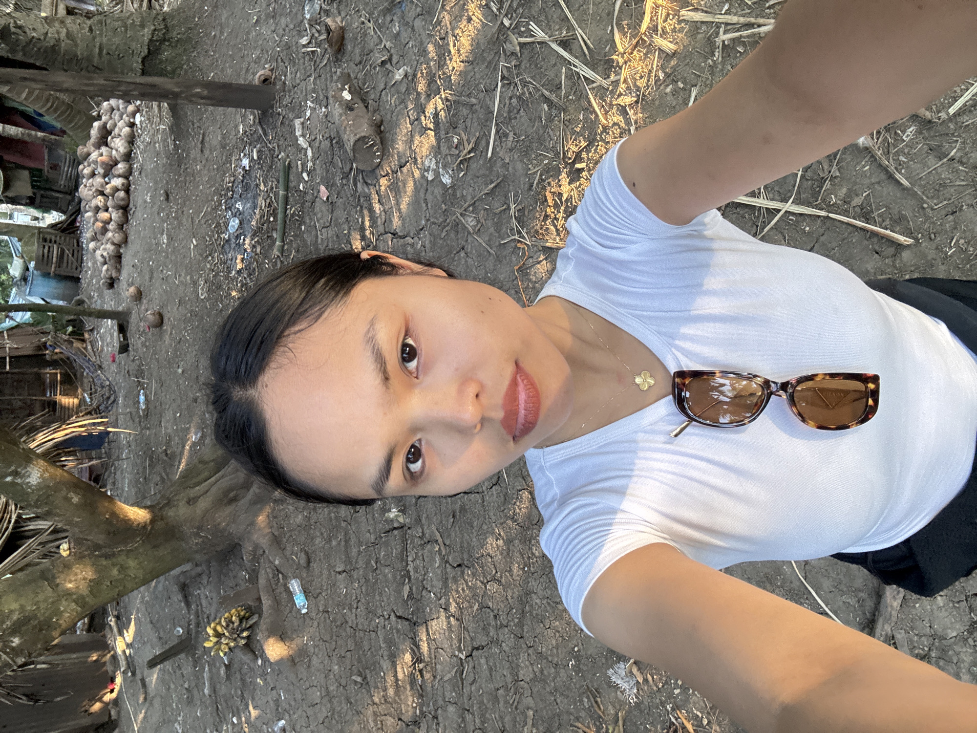 A person, presumably female, is taking a selfie outdoors in Kiblawan, Philippines. She has dark hair pulled back, is wearing a white t-shirt and a gold necklace with a four-leaf clover pendant. A pair of brown tortoise-shell sunglasses rests on her chest. The background consists of dry, cracked earth, vegetation including what appear to be coconut palms and a pile of coconuts in the background. The light suggests it may be late afternoon or early morning. There is some trash scattered around.