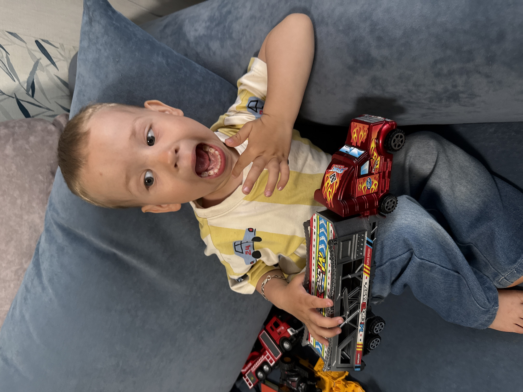 A young child with blond hair lies on a blue couch. The child wears a yellow and white striped t-shirt with a blue car graphic. The child's mouth is open in a wide expression, and their eyes are wide. Several toy vehicles are positioned around the child. These include a red truck with flame decals, a gray car carrier trailer, a red fire truck, and a yellow toy. The child is wearing jeans. The location is not specified in the image, and therefore the city of Rusanovo, Russia, cannot be confirmed.