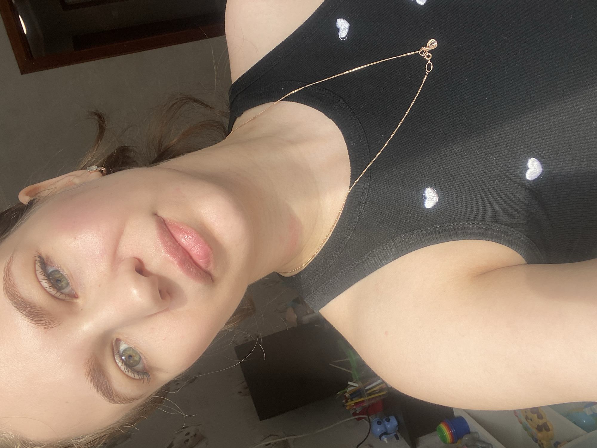 The image is a close-up, chest-up portrait of a fair-skinned individual, likely female, looking directly at the camera. The subject has light green eyes and light brown eyebrows, with what appears to be natural makeup including a light blush and pink lip gloss. Hair is pulled back, with a few wisps visible near the face. She is wearing a black ribbed tank top adorned with small white embroidered heart patterns. A thin gold-colored chain necklace with a small, indistinct pendant is visible around her neck, along with a faint reddish mark on the left side of her neck. A light-colored stud earring is present in her visible right earlobe.
The background, an indoor residential setting, is out of focus and appears dimly lit, suggesting available light source is primarily illuminating the subject from the left. On the left side of the frame, a shelf or desk is partially visible, holding a blue desktop pencil sharpener shaped like a creature, a container of various colored pencils or pens, and rainbow-colored stacking cups. On the right, a dark wooden door frame is discernible. The specific location within the city of Chaikovsky, Russia, cannot be determined from the visual information provided.