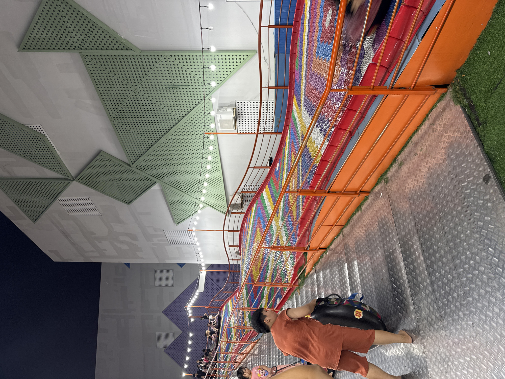 A person stands on a metal platform near the edge of a multi-colored wavy structure with orange railings. The person wears an orange shirt and shorts and carries a black tube. The platform is next to green artificial turf. In the background are a building with a geometrically-patterned ceiling and a night sky.
Based on the style of the wavy structure and other details, this is likely an image taken at the Helio Center amusement park in Đà Nẵng, Vietnam.