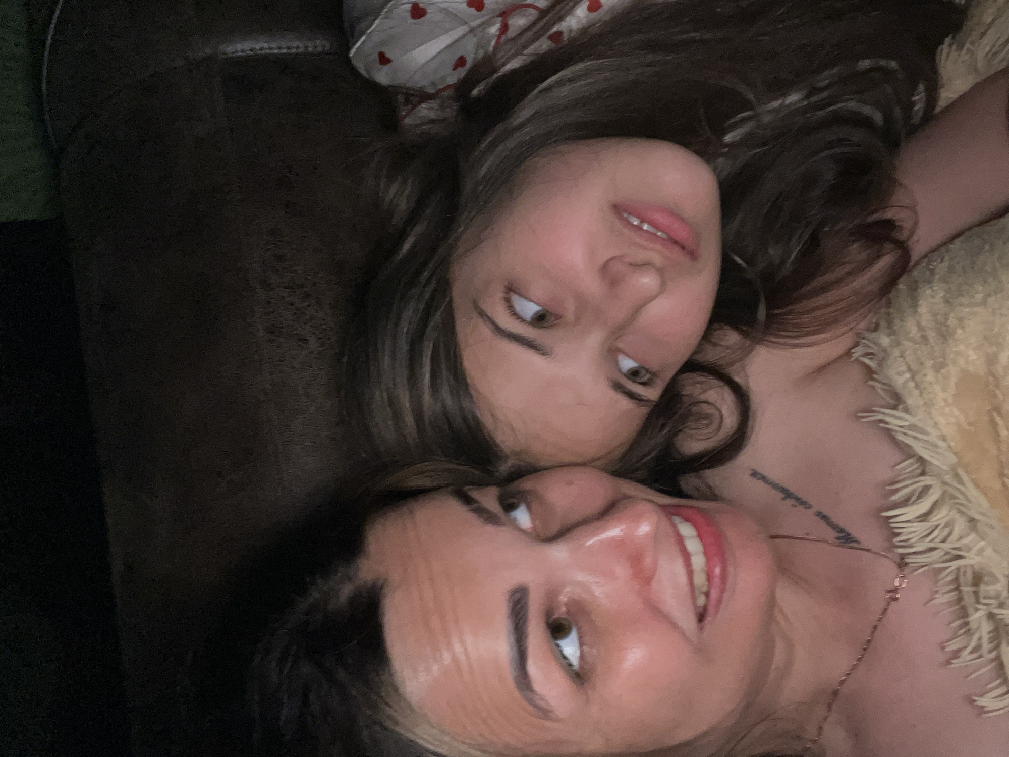 Two females, one adult and one juvenile, are lying down, with their faces close together. The adult is on the left, with light brown hair and fair skin. She is smiling and looking slightly up and to her left. She has a tattoo on her collarbone. The juvenile is on the right, with dark brown hair and fair skin. She is looking to her right. Both are partially covered by a cream-colored, fuzzy blanket. The background is dark and indistinct, suggesting an indoor environment, possibly a couch or bed.