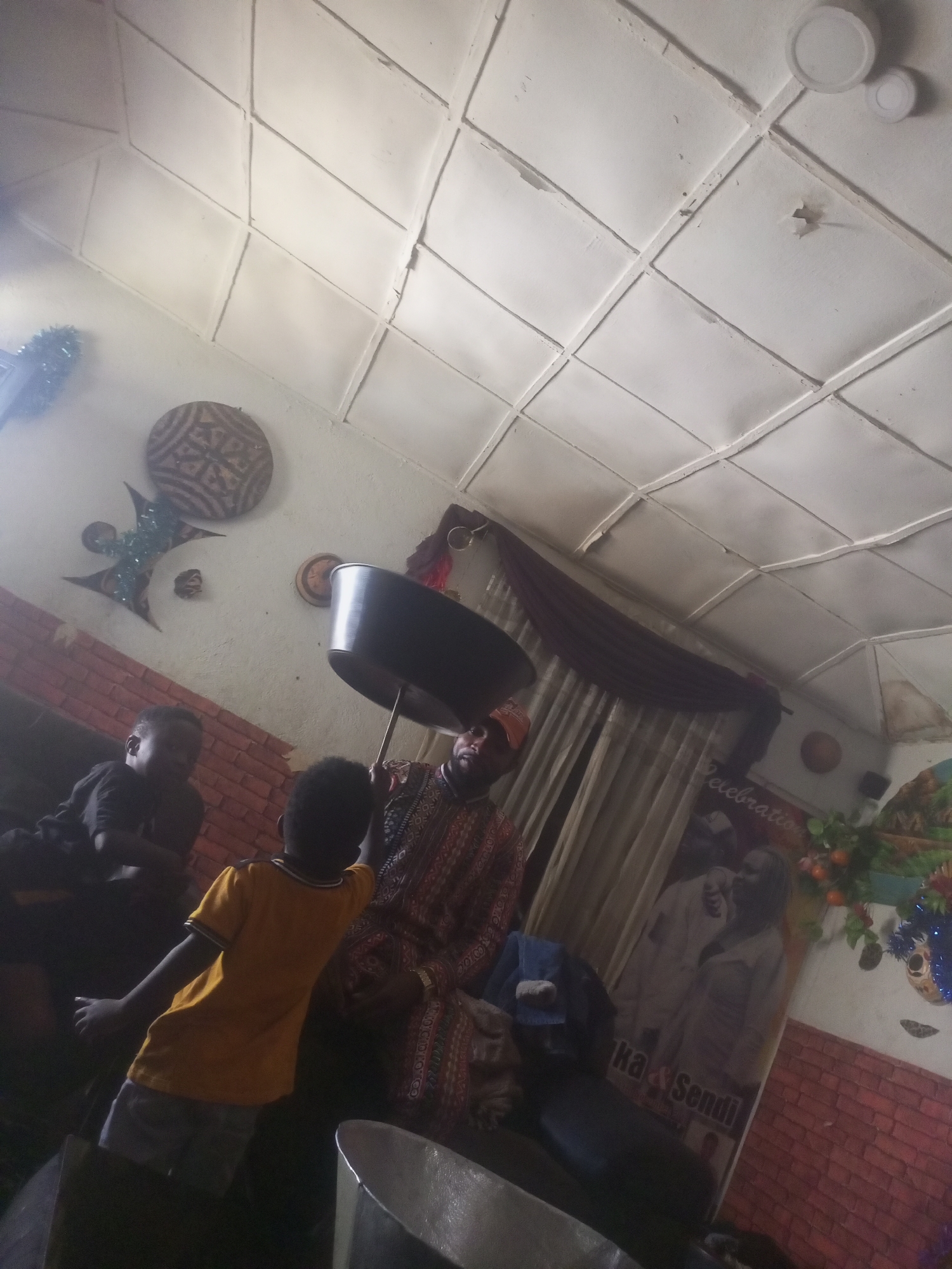 A group of people, including children, are gathered indoors. A man in the foreground is holding up a large metal basin. Another person, a child wearing a yellow shirt and gray shorts, stands in front of him. A man in a patterned shirt is also visible. The room has a tiled ceiling and brick walls, decorated with cultural artifacts and a banner that appears to read "Celebration" and "Ka & Sendi". The lighting is dim.