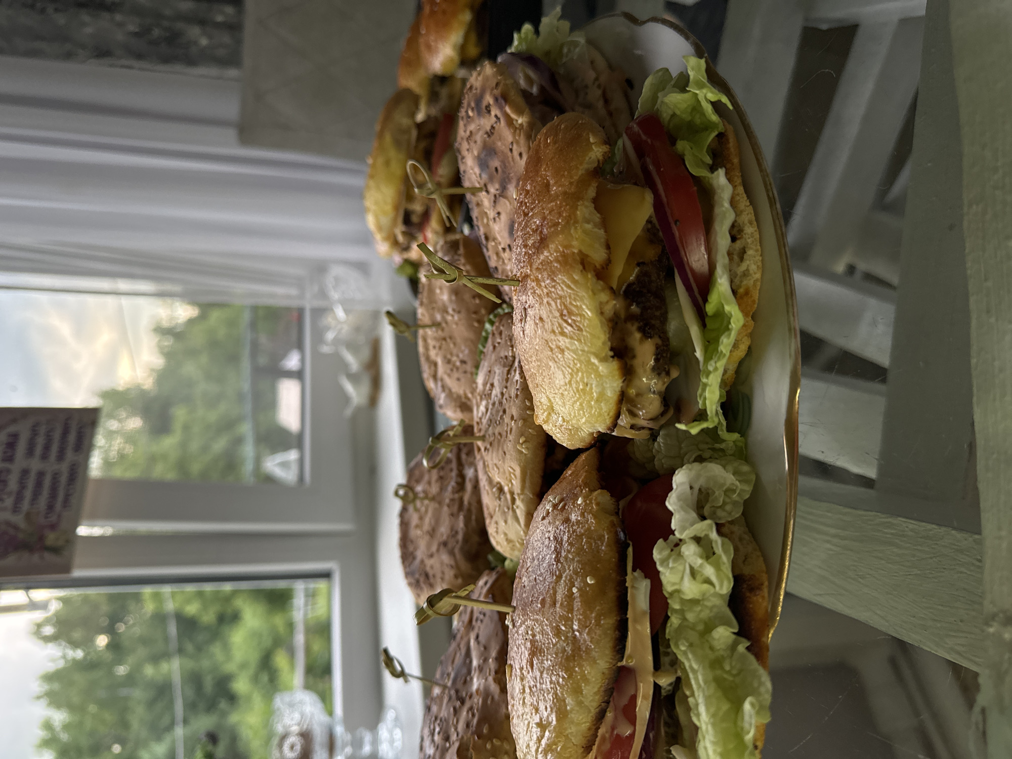 A plate of sliders sits on a reflective surface, likely a table, in front of a window. The sliders are arranged in a line, with several of them stacked in front of others. Each slider appears to have a sesame seed bun, a meat patty, cheese, lettuce, tomato, and red onion. Small wooden skewers are used to hold the sliders together. The window behind the sliders shows a blurry view of green trees and a cloudy sky, suggesting it is daytime with potentially overcast weather. The scene appears to be indoors, with the light suggesting natural light from the window. There are some faint scratches or marks on the reflective surface. No people are visible in the image.