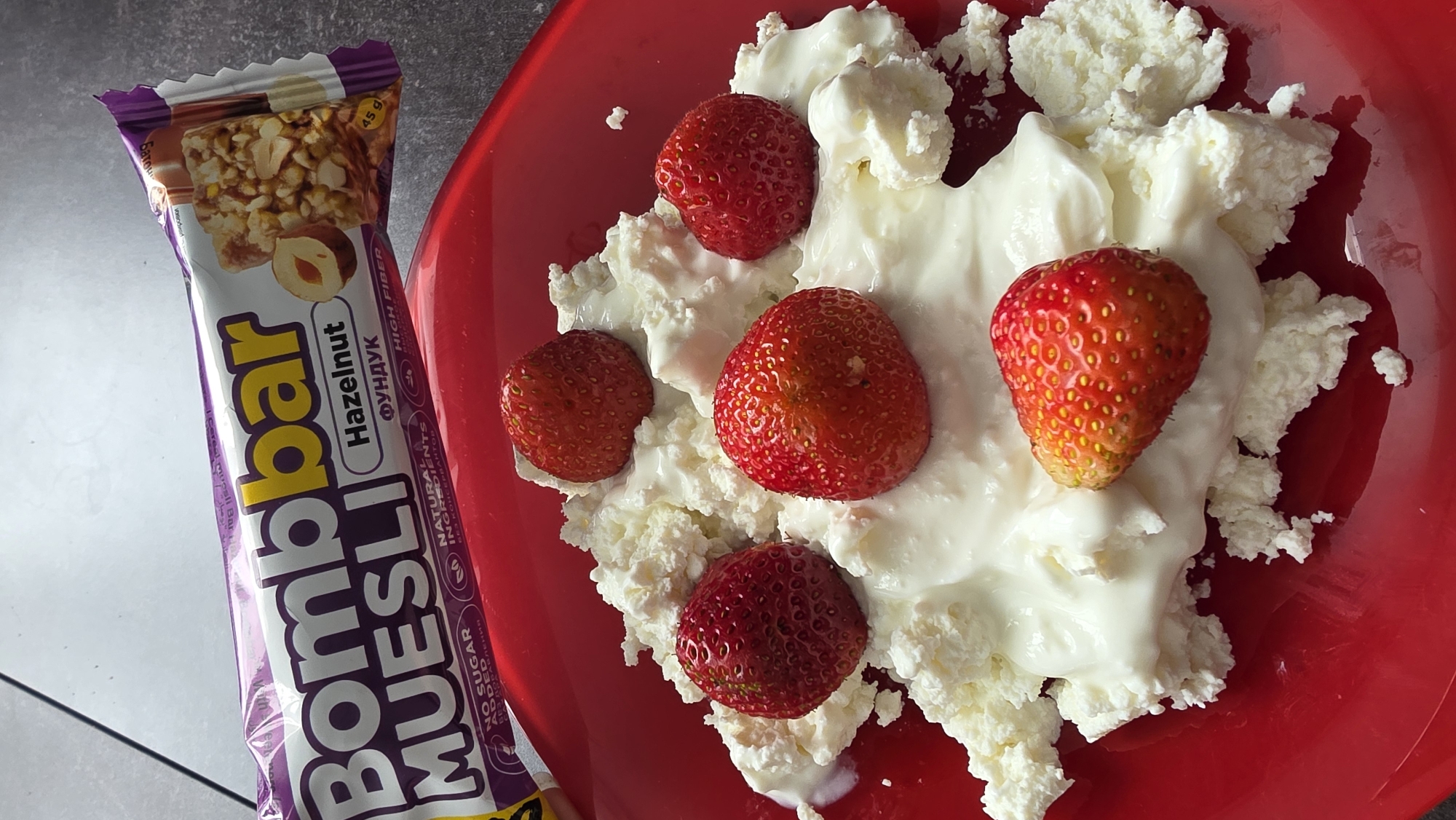 A Bombbar Muesli Hazelnut cereal bar is placed diagonally across the top of the image, partially over a bright red plate. The plate contains a generous serving of white, crumbly cottage cheese topped with a dollop of yogurt. Five fresh strawberries are scattered on top of the cottage cheese and yogurt. One strawberry is whole and bright red, positioned in the upper right quadrant of the plate. Another whole, bright red strawberry is in the upper left quadrant. A third whole, red strawberry is in the center of the plate, slightly to the left of center. A fourth whole, red strawberry is in the lower right quadrant, and a fifth strawberry, partially sliced and showing orange hues, is in the lower center of the plate. The background is a dark, textured surface, possibly a table. The scene appears to be indoors, with overhead lighting.